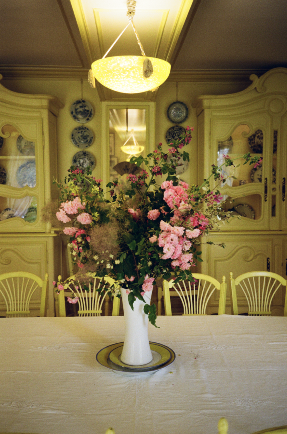Une grande vase blanche avec un bouquet de fleurs roses, sur une nappe blanche, dans une salle avec des chaises jaunes et un buffet ancien en arrière-plan.