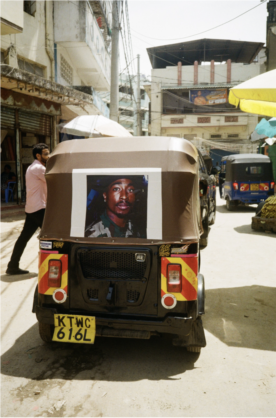 Une auto-rickshaw avec une photo d'un homme en uniforme militaire collée à l'arrière, circulant dans une rue animée avec d'autres véhicules et un homme marchant à côté. L'environnement montre des bâtiments en ancien état et des stands de marché.