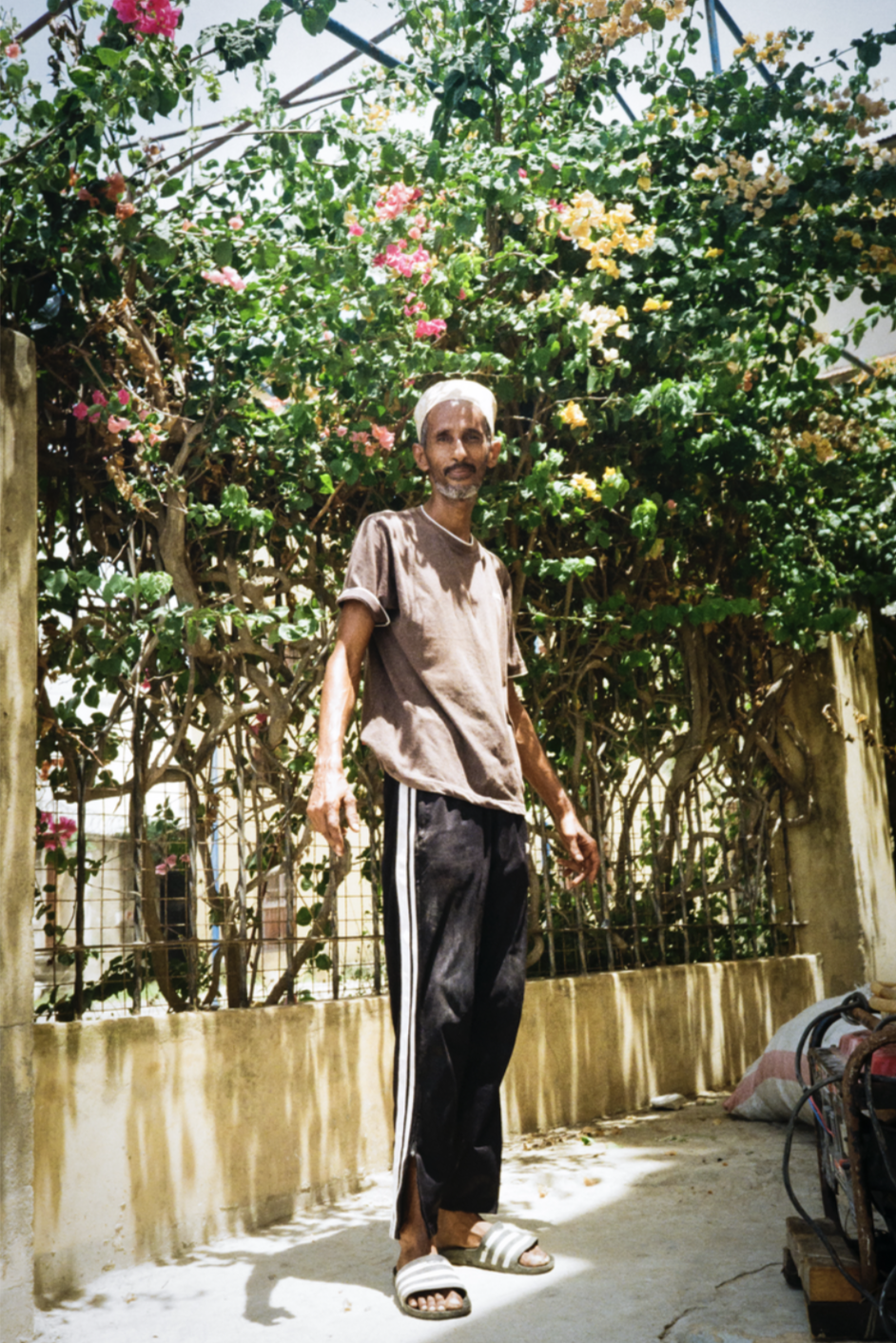 Un homme debout devant une clôture avec des arbres et des fleurs colorées en arrière-plan, portant un t-shirt beige, un pantalon noir avec des bandes blanches sur les côtés, des chaussons et un turban blanc.
