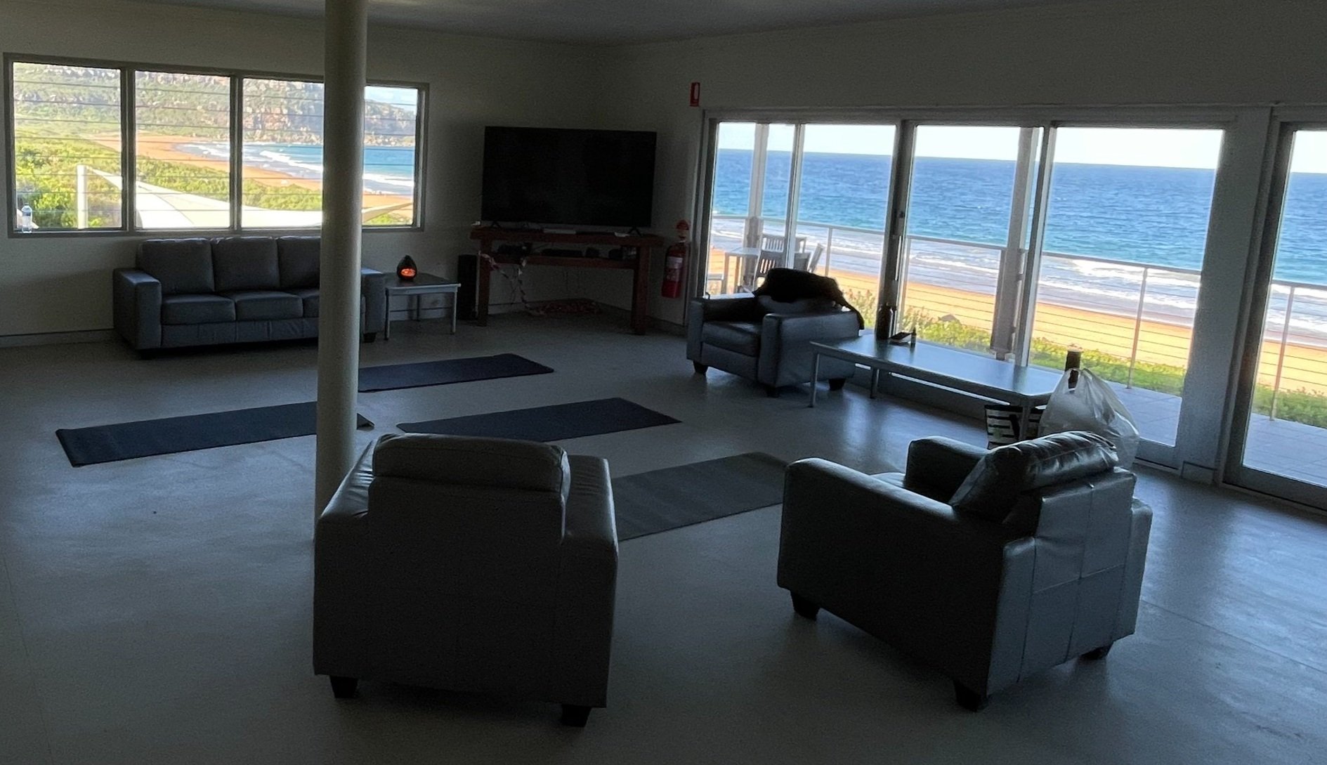 Living room with large windows and sliding glass doors overlooking a beach and ocean, contains leather sofas, a TV, and yoga mats on the floor.
