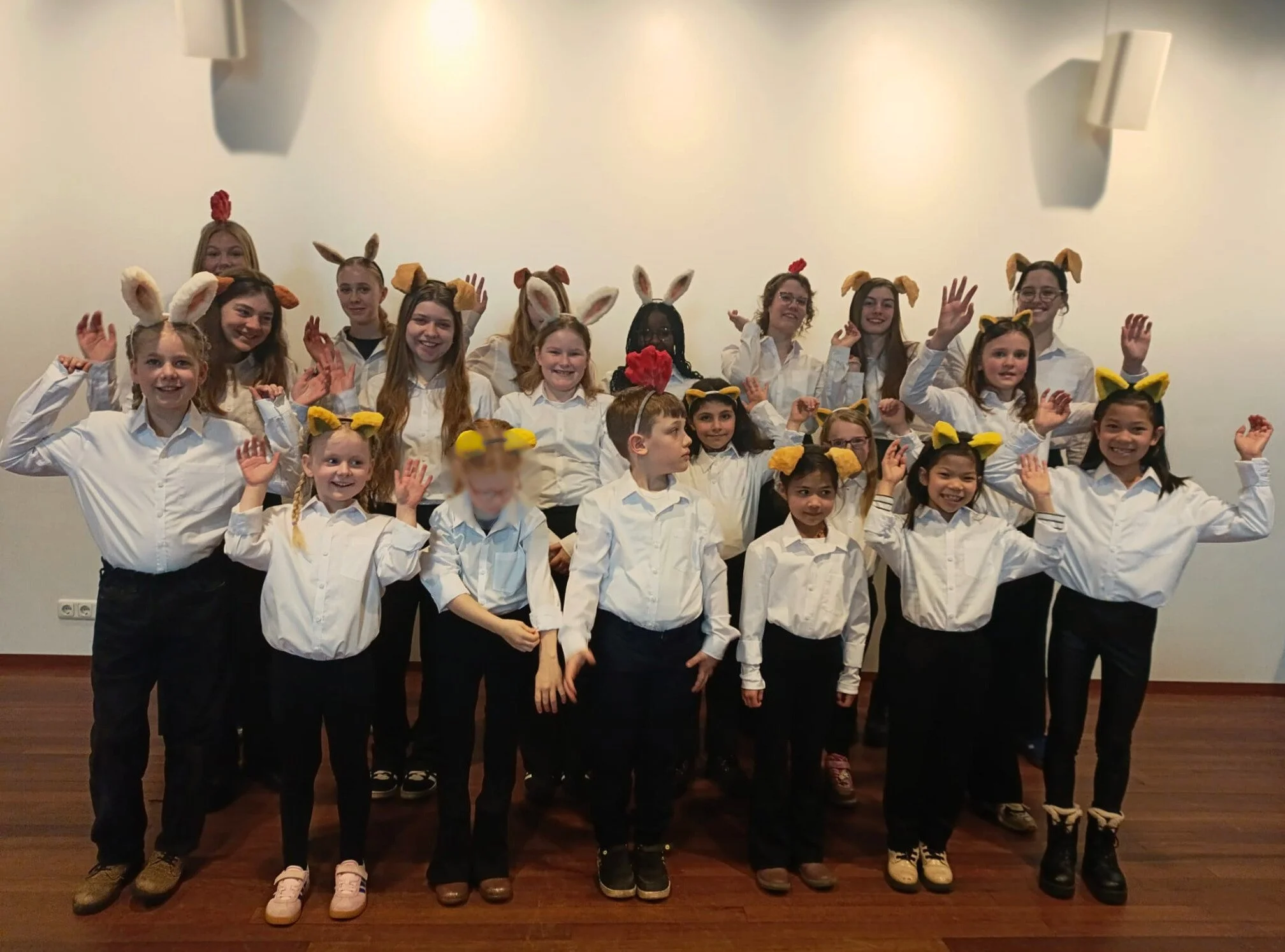 Groep kinderen in witte shirts dragen dierenoren en hoorns, staan op een houten vloer, poseert voor een witte muur; sommigen zwaaien of maken handgebaren.