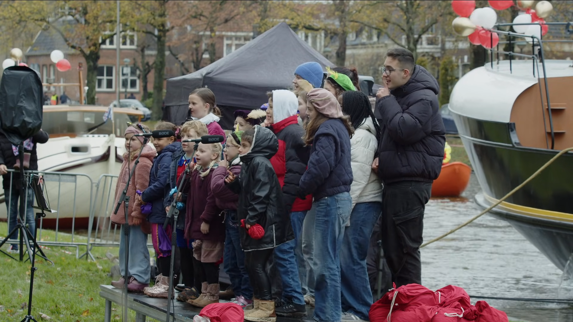 Groep mensen op een podium bij een water, met boten op de achtergrond, tijdens een buitengebeurtenis in de herfst.