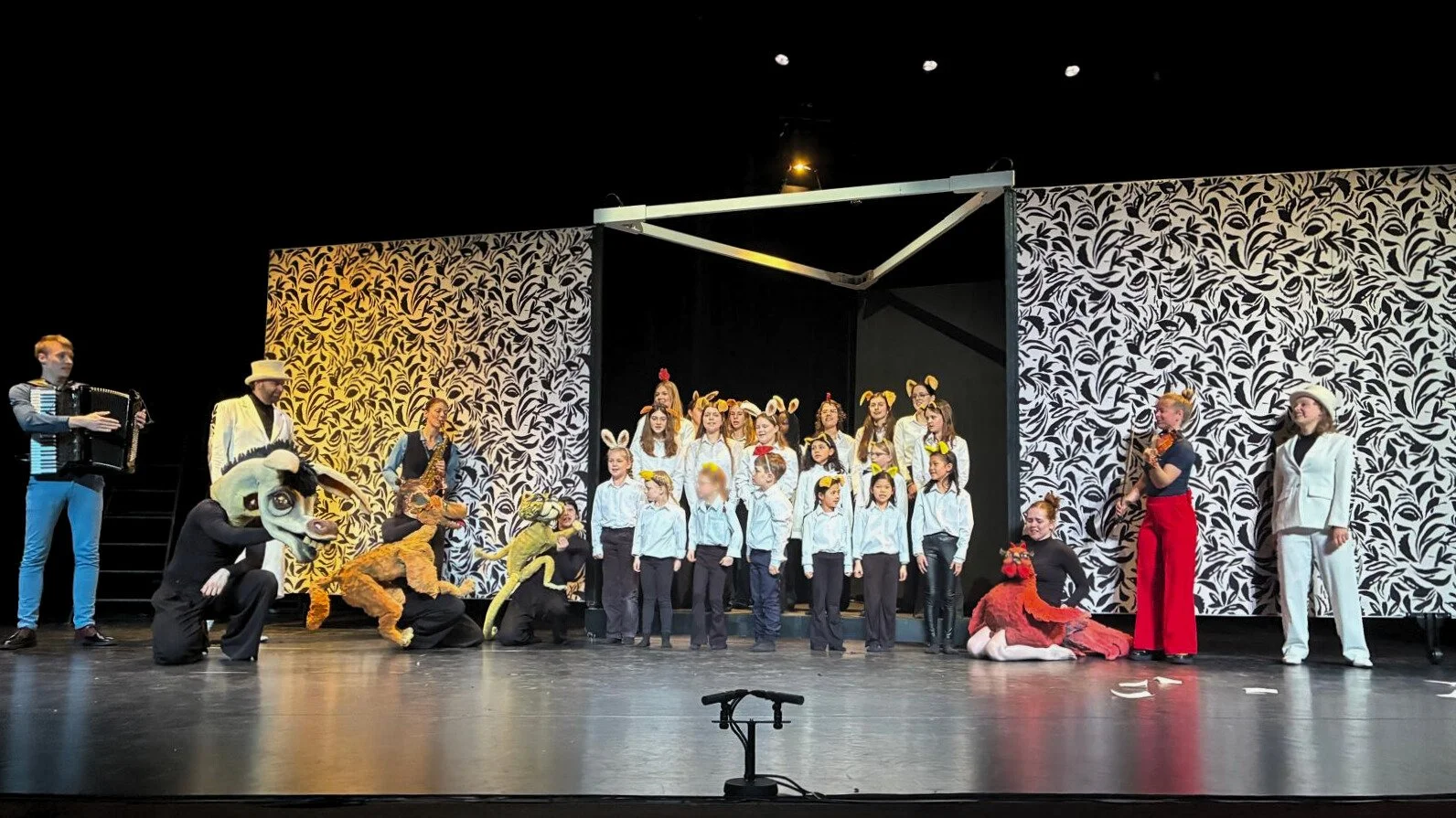 Theatergroep met kinderen en volwassenen in dierenkostuums tijdens een voorstellings op een toneel met zwart-wit bladermotief achterwand.