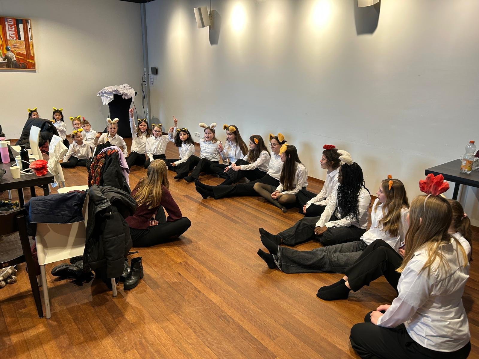 Groep kinderen verkleed als dieren, zittend op de vloer tijdens een repetitie of voorstelling in een kamer met houten vloer.