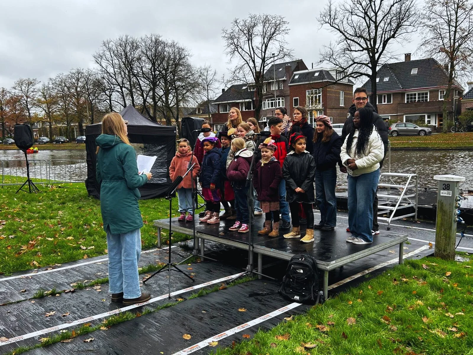 Mensen, waaronder kinderen en volwassenen, staan op een klein podium bij een rivier, terwijl een vrouw met een microfoon spreekt. Het lijkt een buitengebeurtenis of optreden in een park, met een nieuwbouw wijk op de achtergrond.