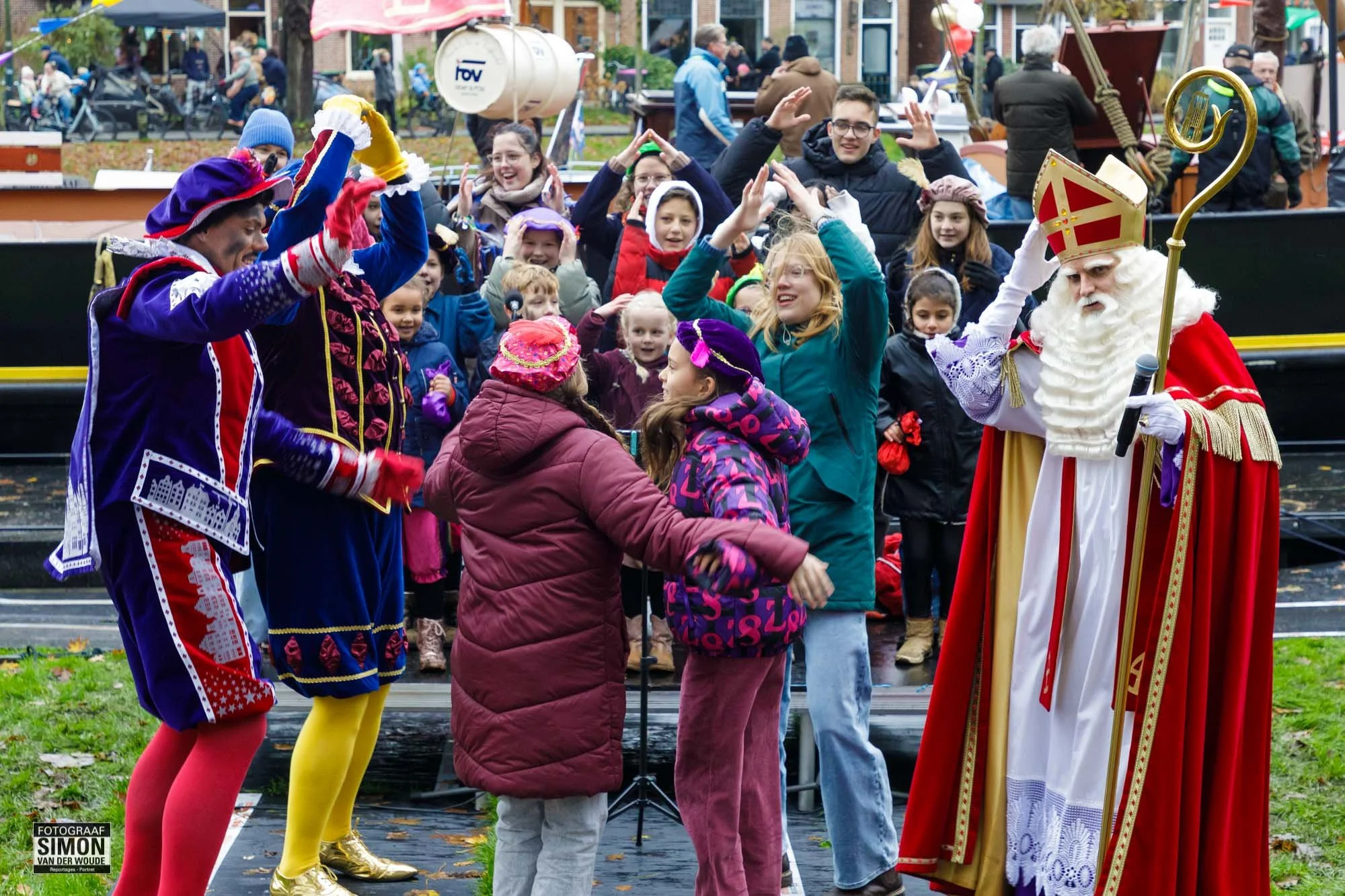Sinterklaas en Zwarte Piet vieren feest met kinderen in een park, met een boot op de achtergrond.