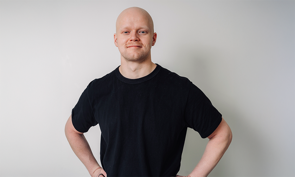 A smiling man with a bald head is outdoors in front of a mountain range during sunset, wearing a dark red shirt.