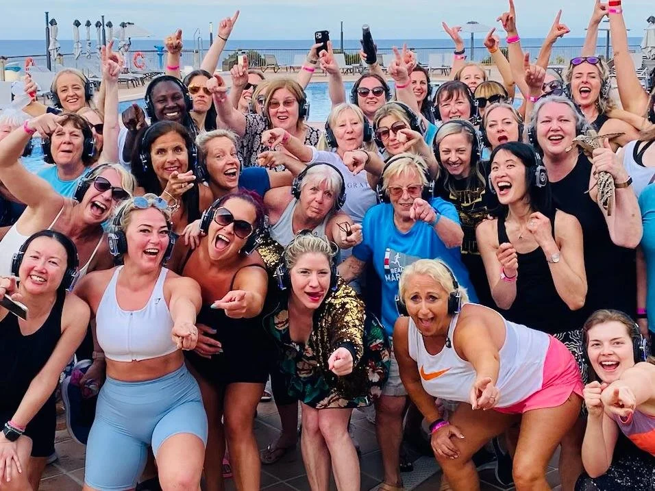 Large group of women cheering and smiling on a cruise ship deck, some wearing headphones, with the ocean in the background.