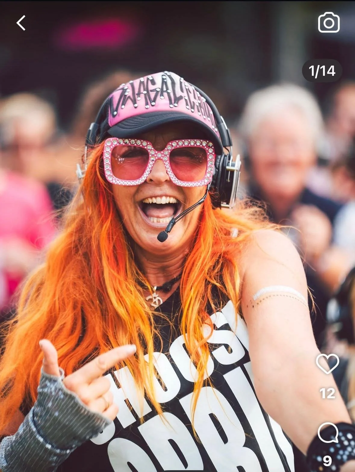 A woman with bright orange hair wearing large pink sunglasses, a black cap, and a headset, smiling and pointing towards the camera at a lively outdoor event.