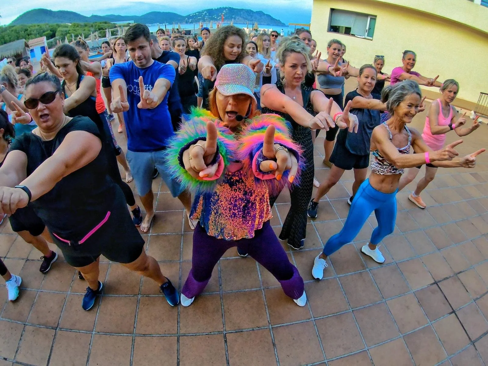 Group of people participating in an outdoor dance class or exercise session during daytime, with a colorful background of mountains, water, and sky.