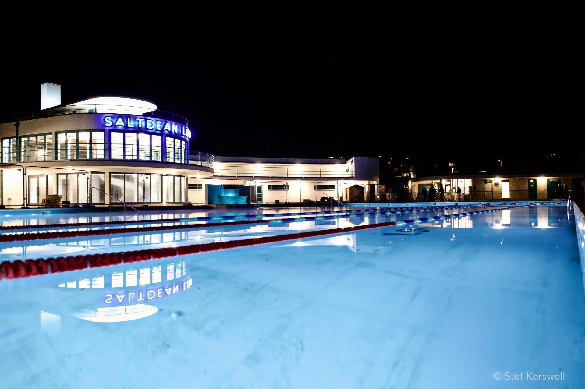Night view of an outdoor swimming pool at the Saltdean Lido, with illuminated building and poolside area reflected in the water.