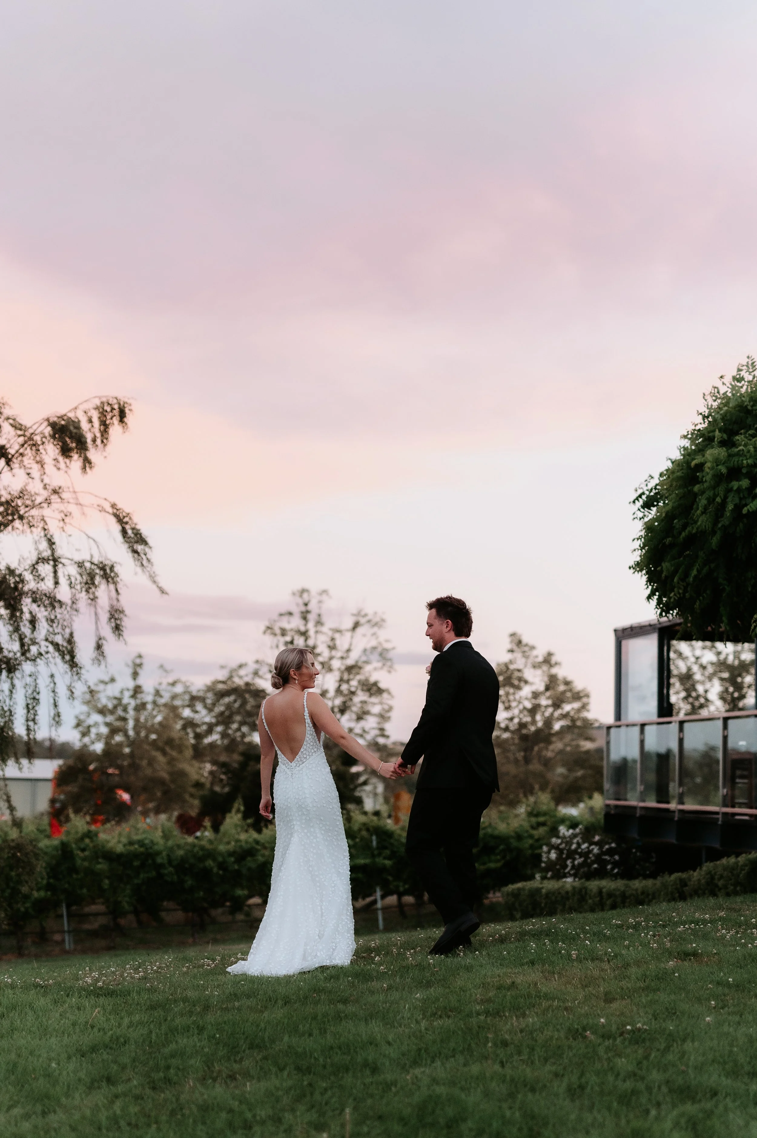 A woman in a white dress holding hands with a man in a black suit. They are looking at each other. The background is a winery with a sky of soft pinks and purples.