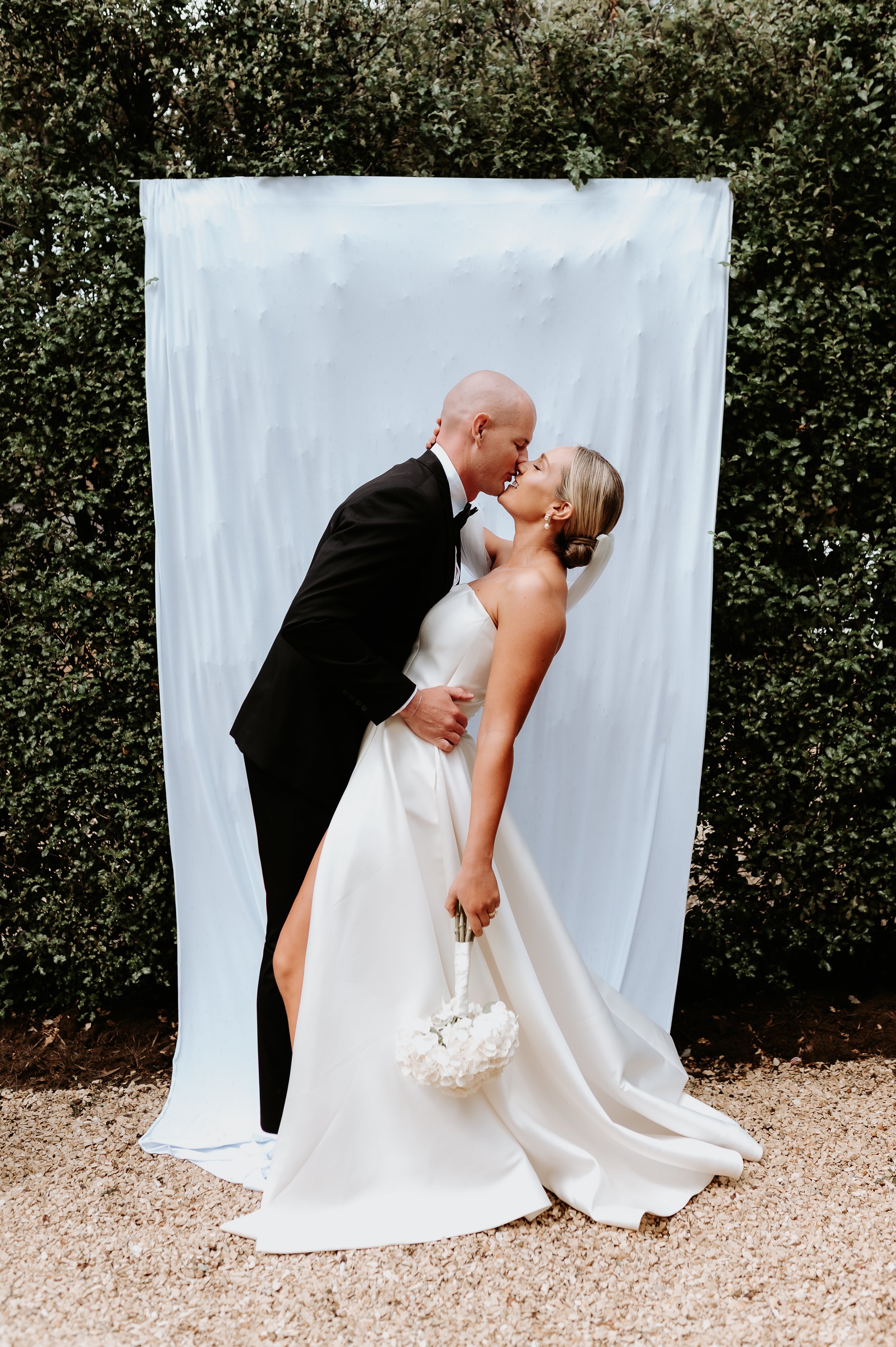 A woman in a white dress and a man in a suit embracing. The background is a green hedge with a white backdrop strung along the greenery.