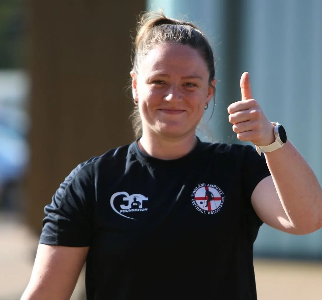 A smiling woman with a ponytail giving a thumbs-up gesture, wearing a black T-shirt with logos, outdoors on a sunny day.