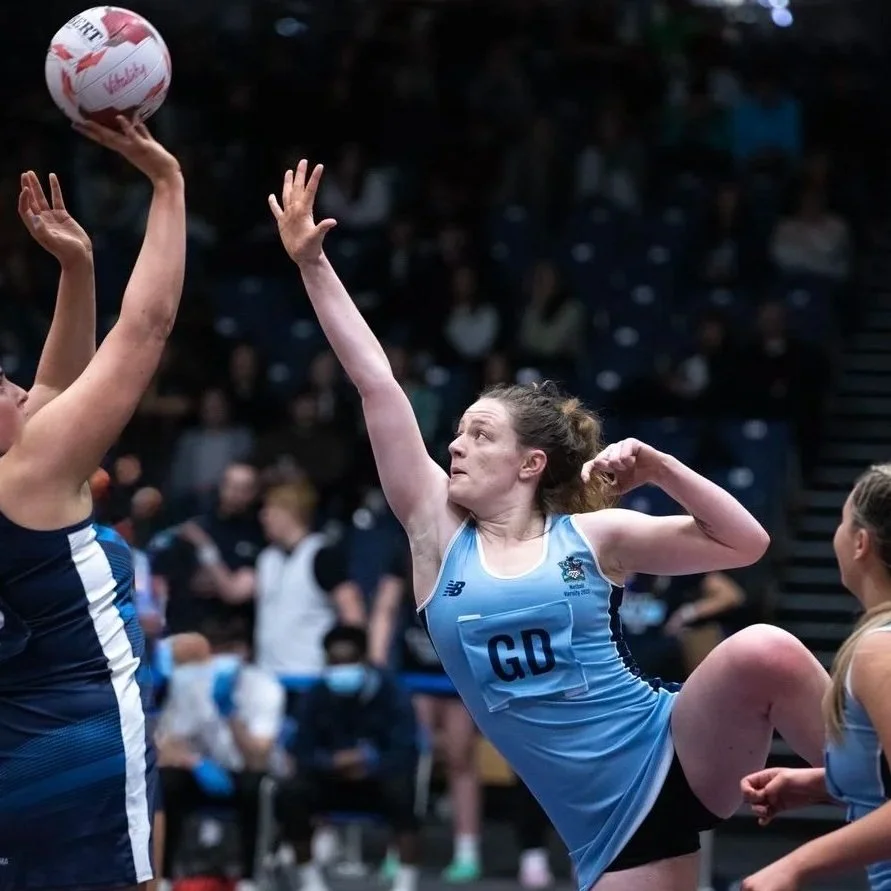 A female netball player in a light blue uniform with 'GD' on her chest, reaching high to block or intercept a ball from an opposing player during a game.