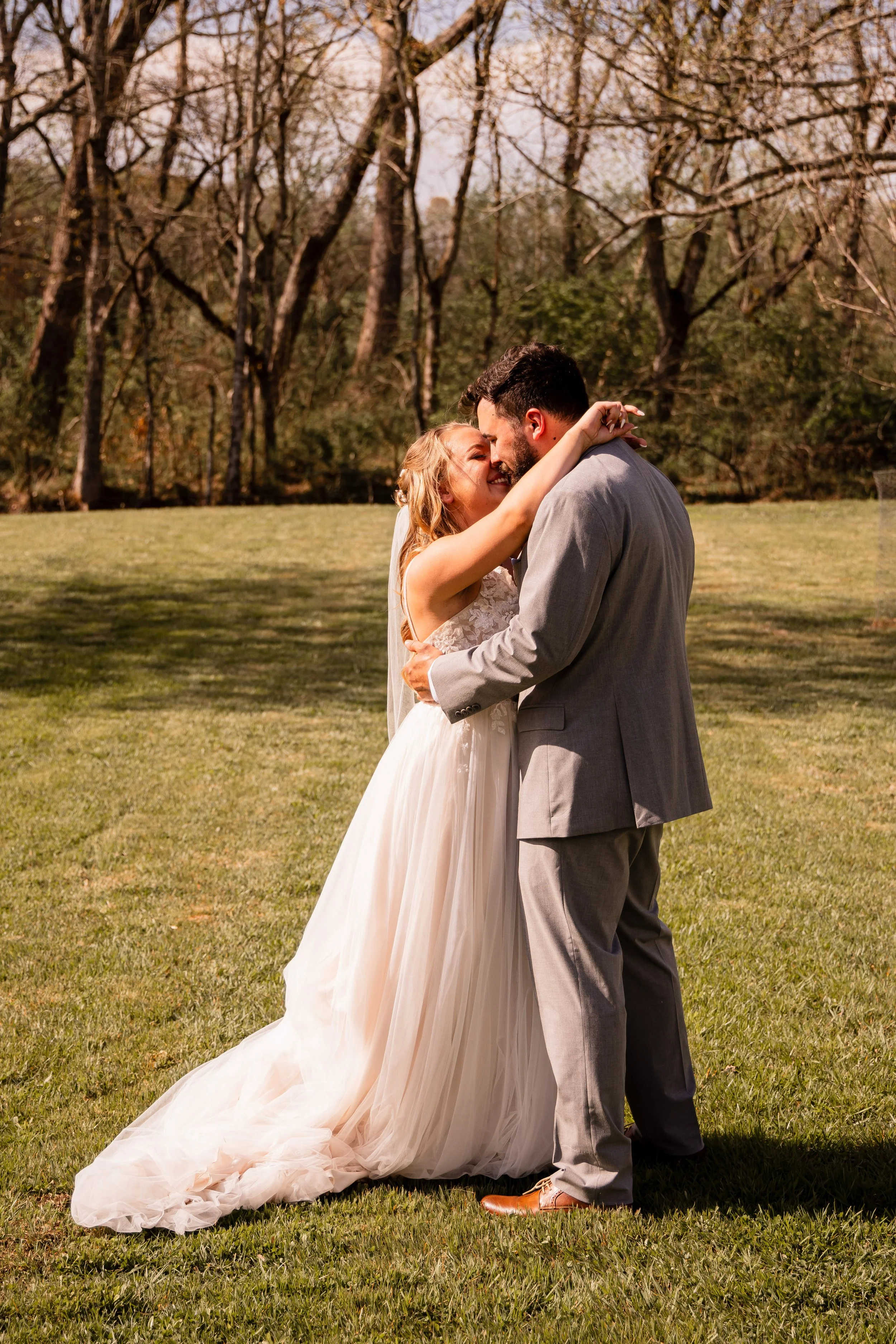 A bride and groom embrace and smile outdoors on their wedding day, with trees in the background.