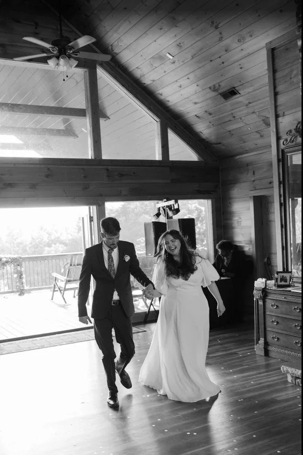 A bride and groom dancing together inside a Gatlinburg cabin with large glass windows and a vaulted ceiling. The bride is wearing a white wedding dress, and the groom is in a dark suit.