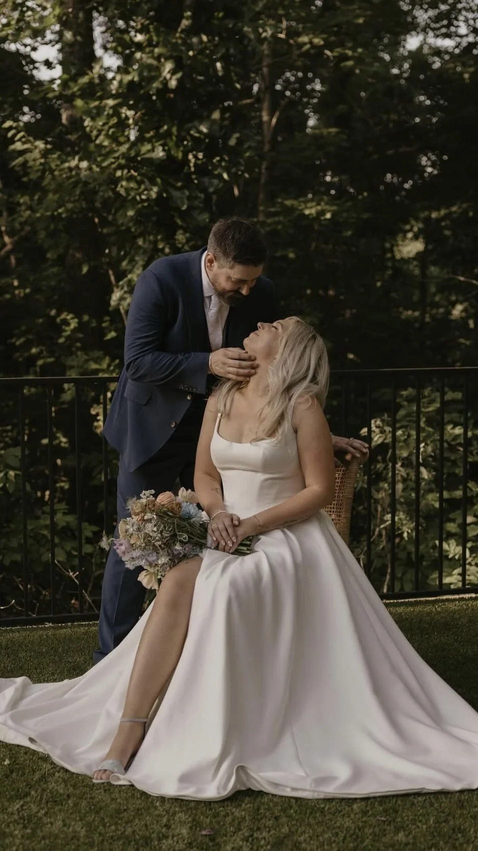 A bride sitting on a chair outdoors, holding a bouquet of flowers, with a man dressed in a suit leaning over her, touching her neck, during a wedding photo shoot.