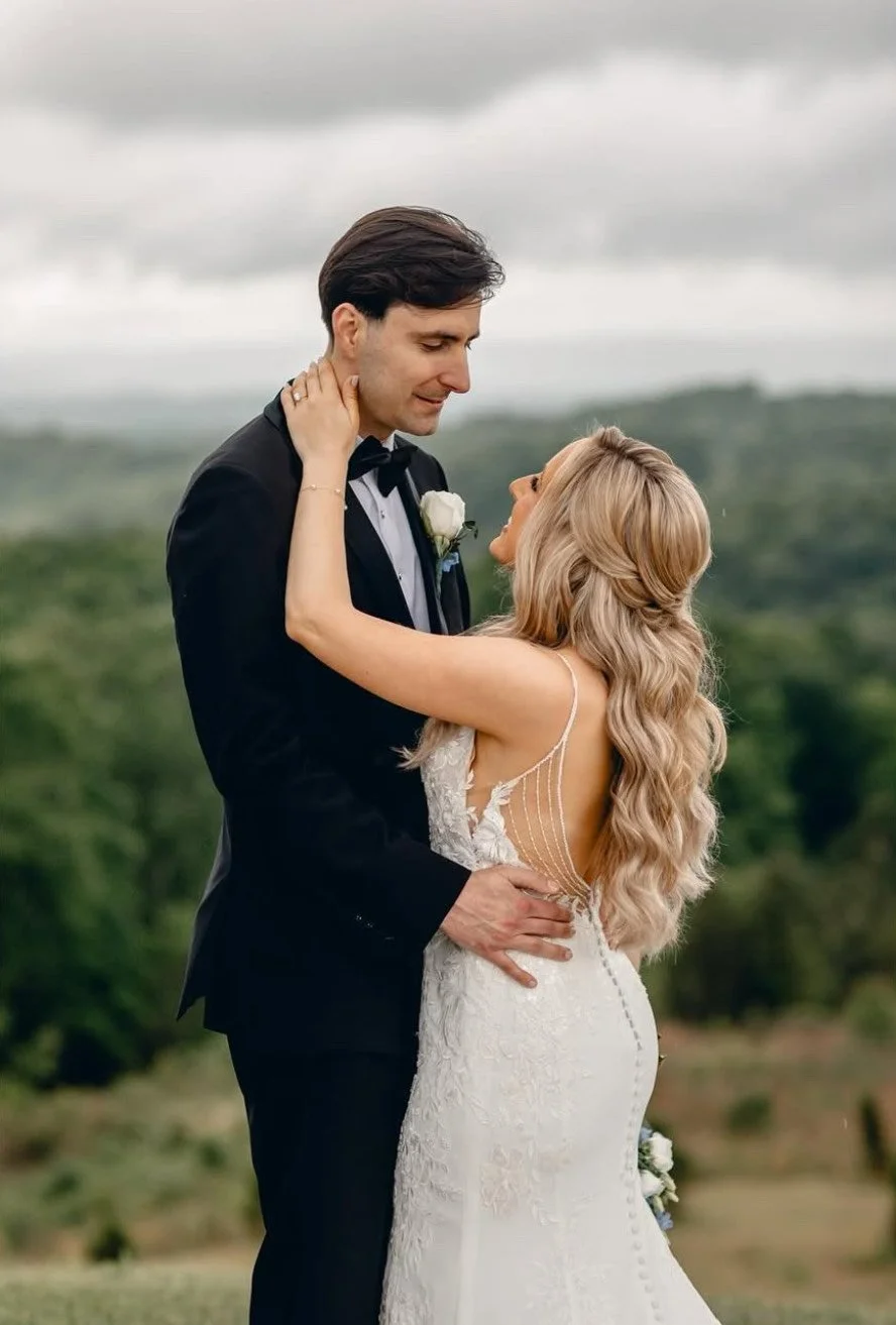 Bride and groom embrace outdoors, with the bride in a white lace dress and the groom in a black tuxedo with a bowtie, holding each other tenderly on a cloudy day.