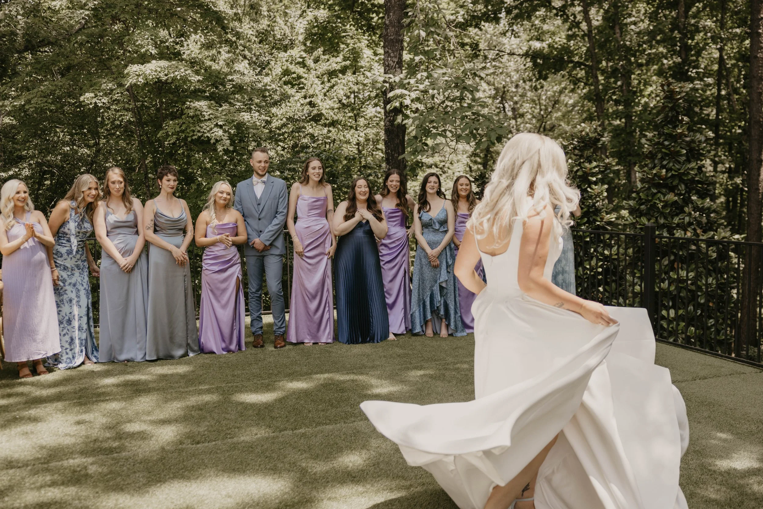 A bride in a white wedding dress twirls outdoors with a group of women in pastel-colored dresses and one man in a suit, standing in a forested area with trees in the background.