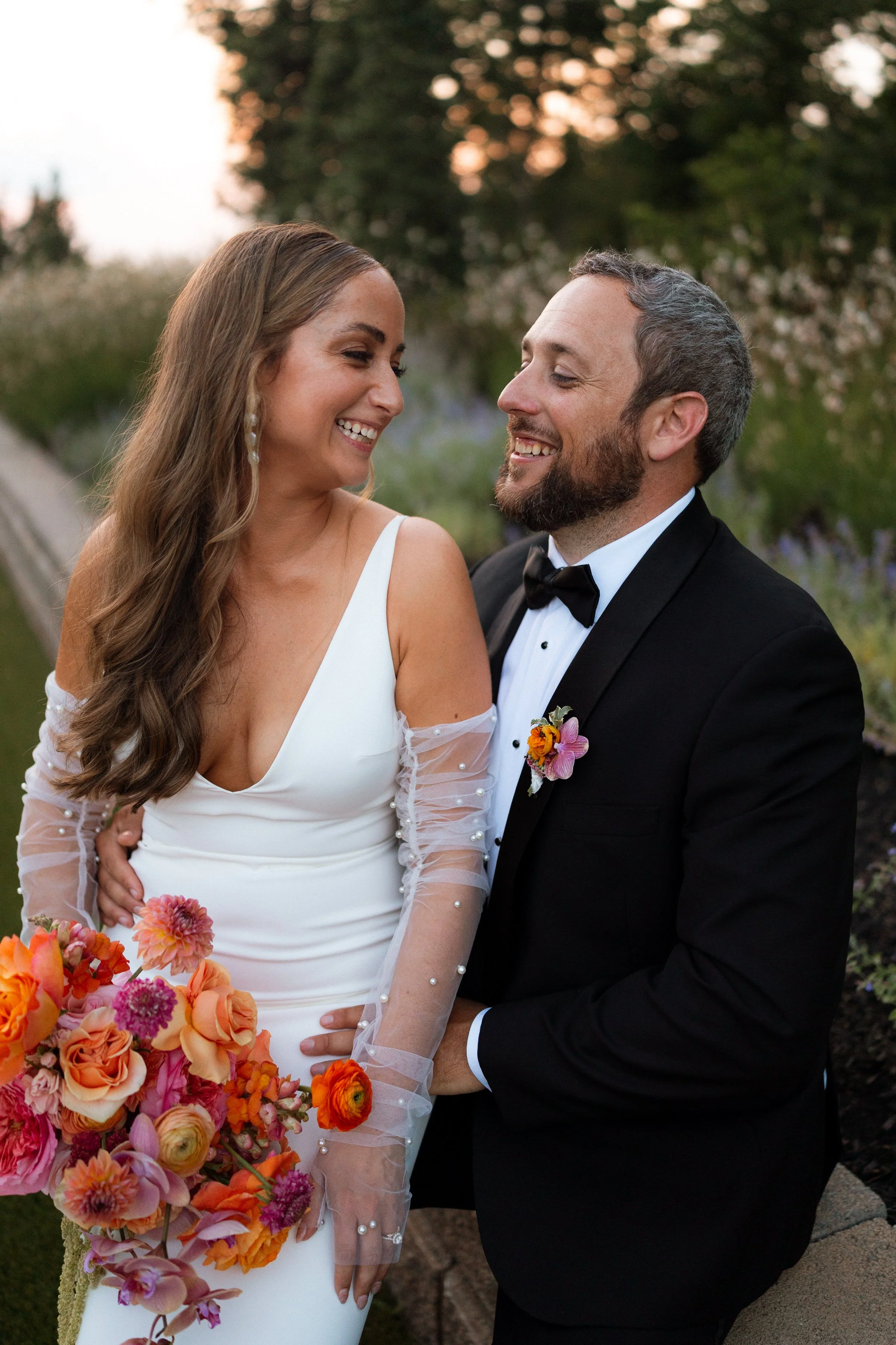 A bride and groom in wedding attire smiling at each other outdoors during sunset, with the bride holding a colorful bouquet of flowers.