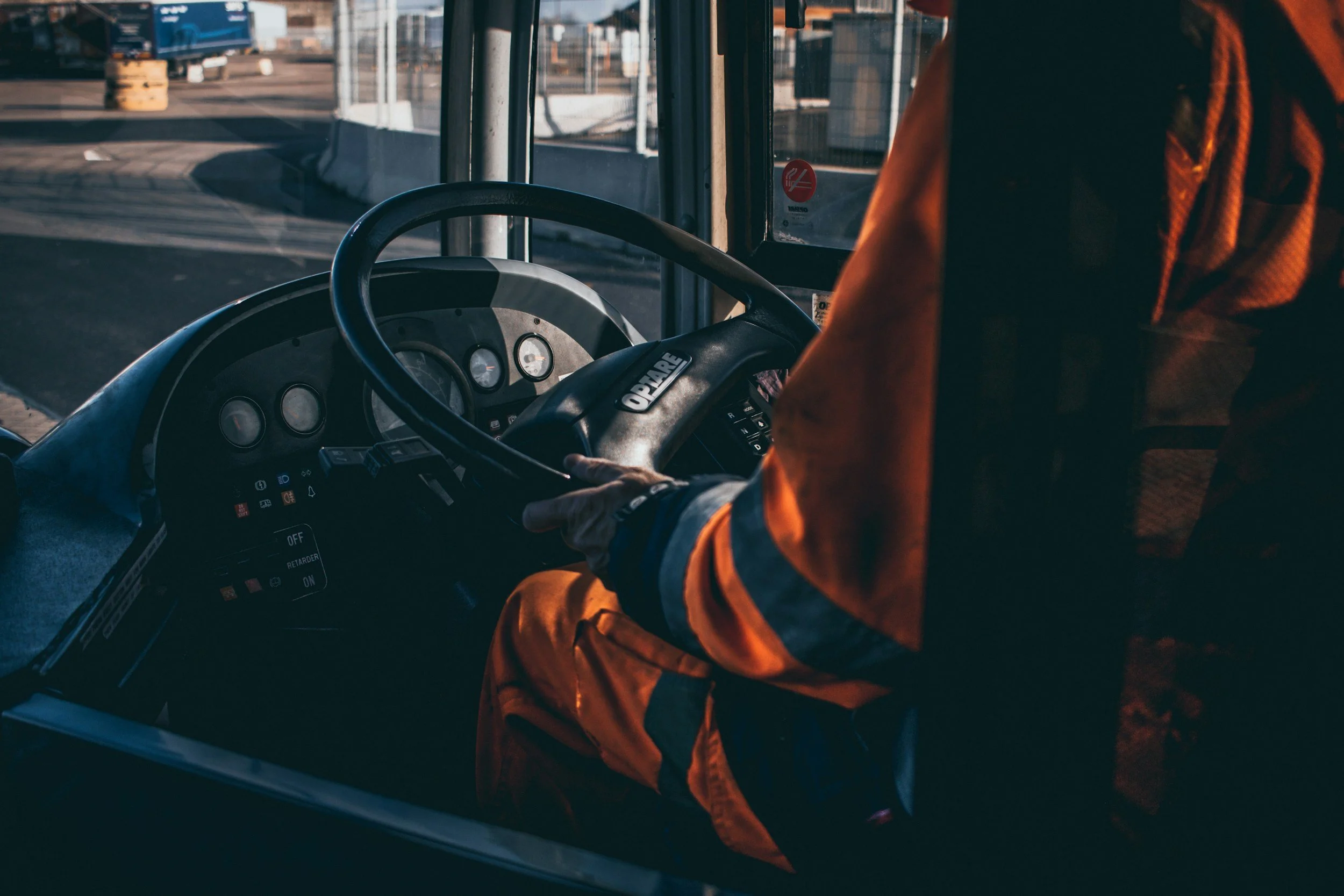 A bus driver with a safety vest operating a bus, with the city street visible through the front window.