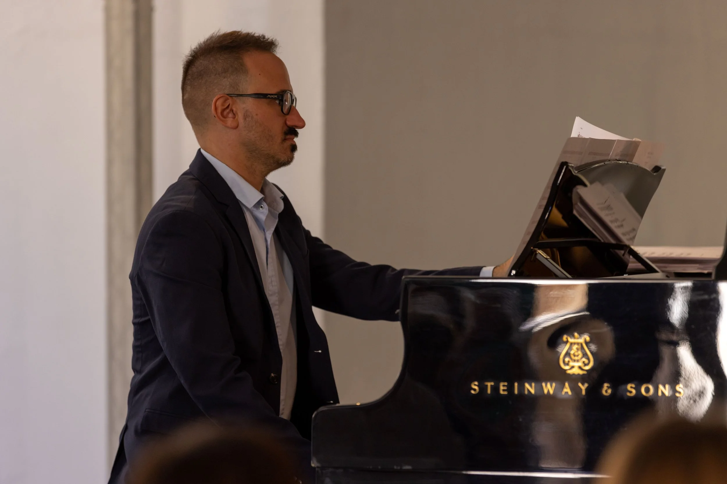 Hombre tocando el piano negro de la marca Steinway & Sons, vestido con saco oscuro y lentes, en un ambiente interior.