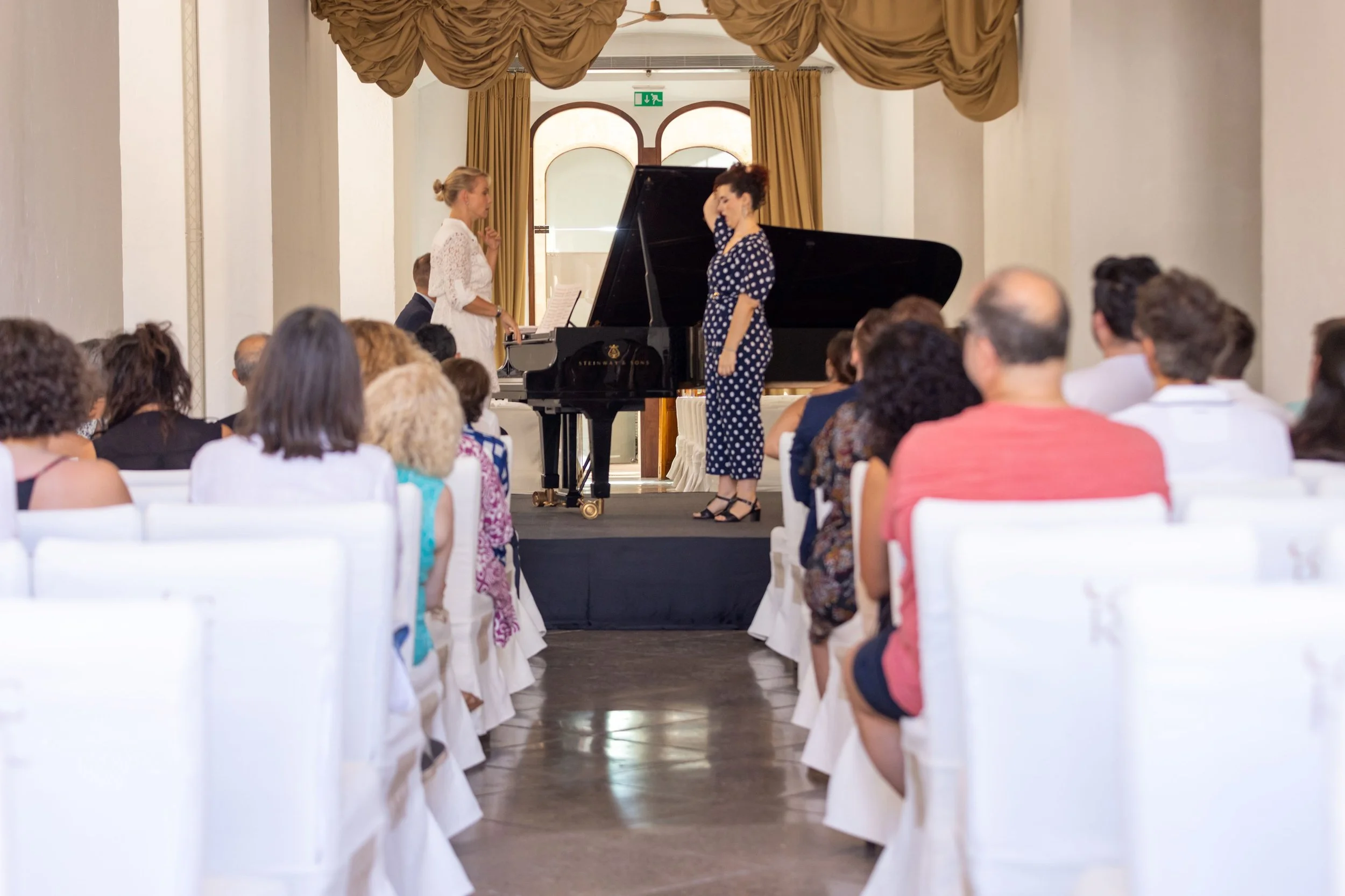Un concierto de piano con una cantante en un escenario, asistido por una audiencia en un salón elegante con cortinas doradas.