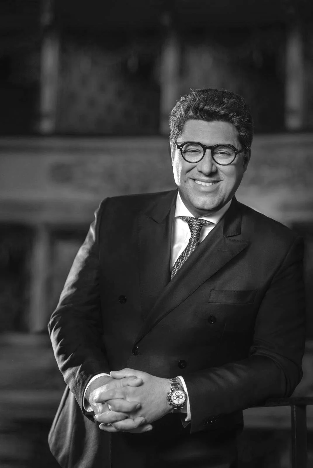 Portrait of Luca Salsi, smiling, wearing glasses and suit, in a wooden setting, black and white.