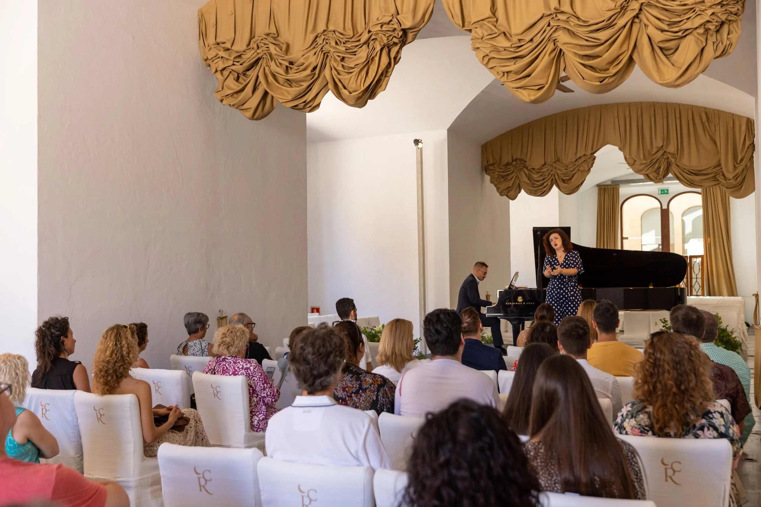 Un concierto de piano con una cantante en un salón elegante con cortinas doradas y un público sentado