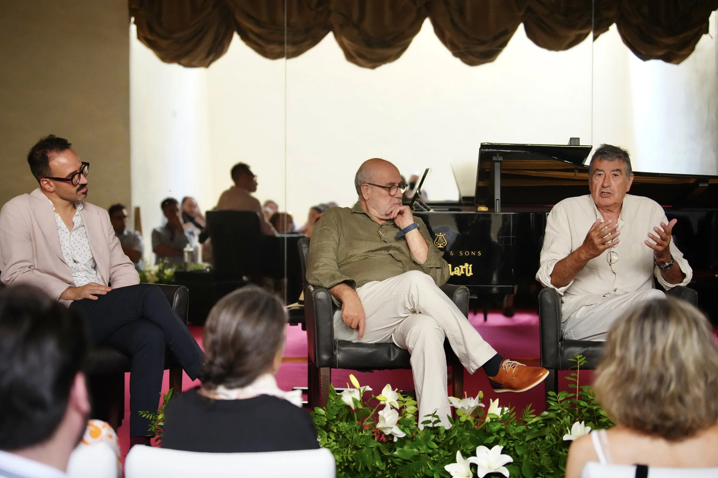 Tres hombres sentados en una mesa en un evento, hablando y escuchando. Detrás hay un piano de cola negro y un grupo de personas en el fondo. Frente a ellos, hay varias personas en la audiencia. La escena tiene plantas y flores en primer plano.