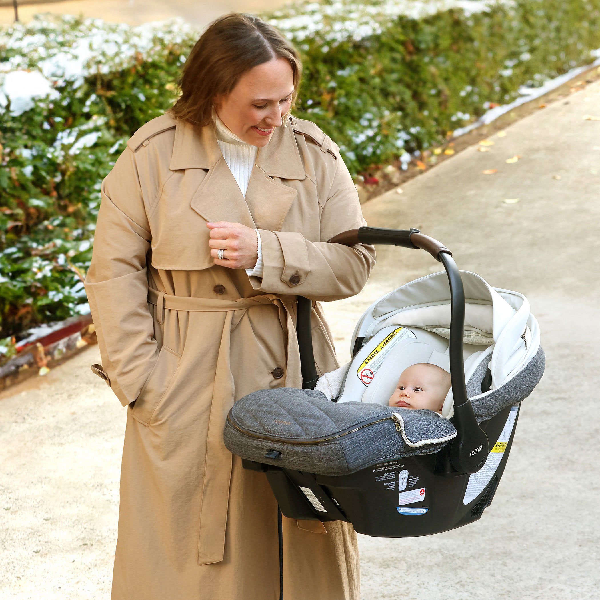 A woman wearing a long coat and carrying a baby in an infant car seat. The car seat has a plush gray cover.
