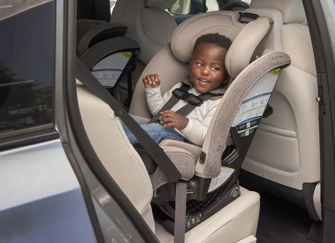 A toddler secured in a rear-facing car seat in the back of a vehicle, looking out the open door and smiling.