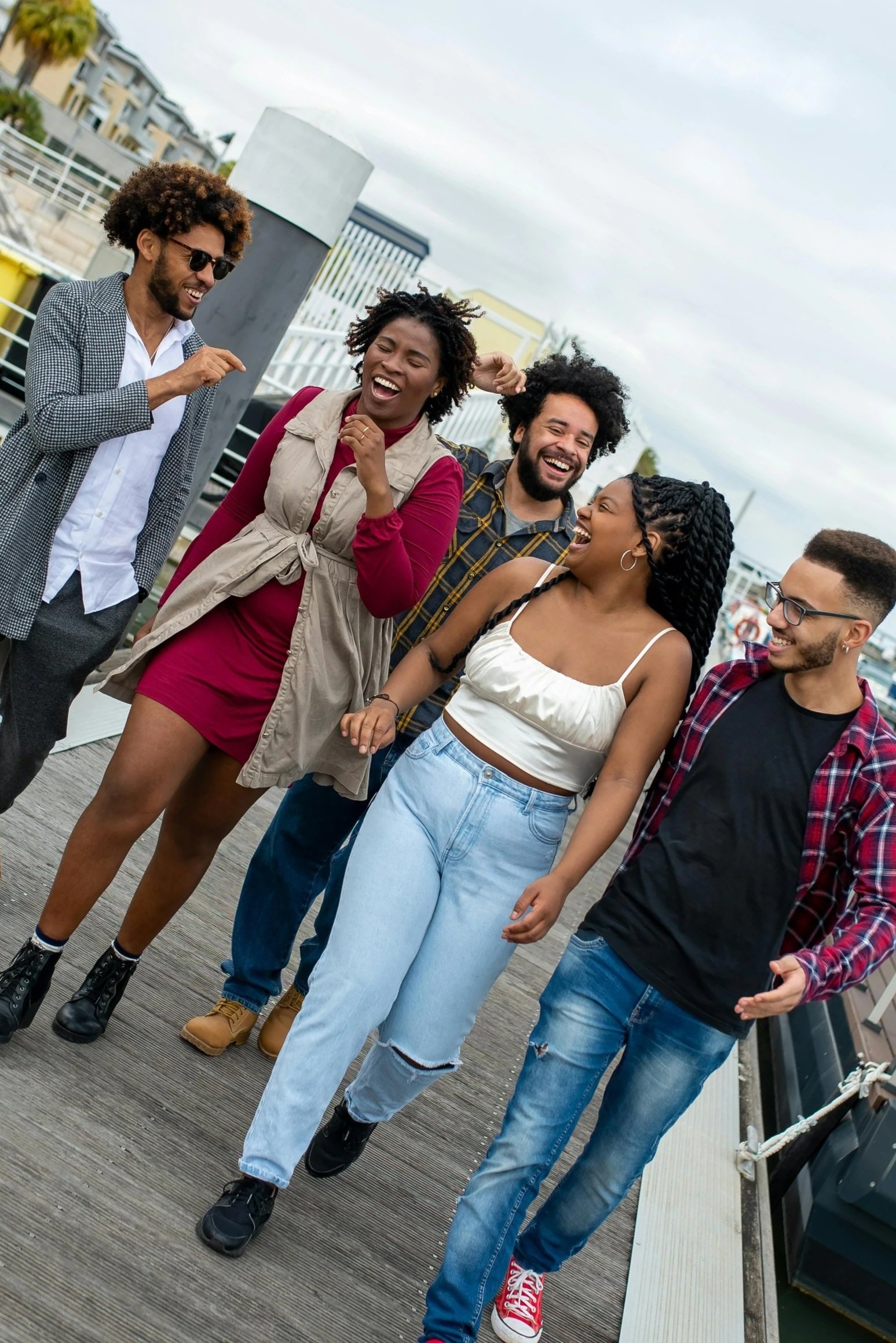 Group of five friends walking and laughing outdoors on a cloudy day, dressed casually with trendy clothes and hairstyles.