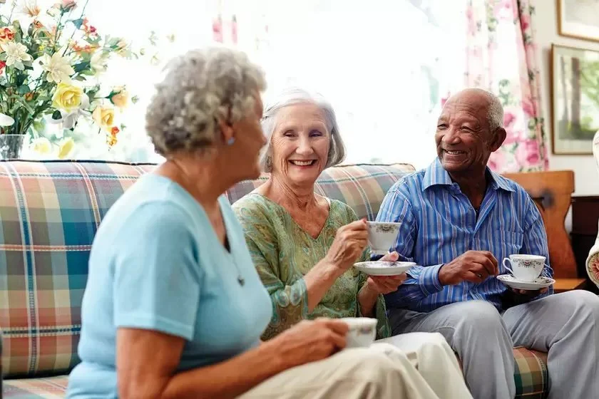 Three elderly people enjoying tea and conversation on a plaid sofa in a brightly lit room with floral curtains and artwork on the wall.