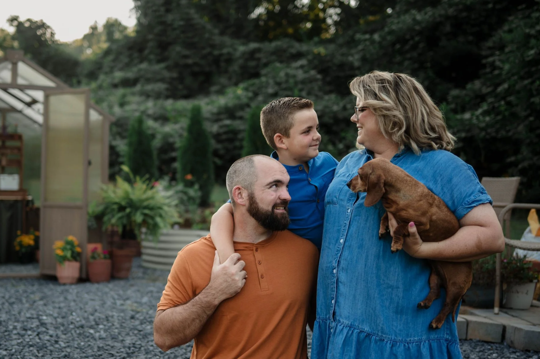 The Broome family with a small dog Rosie  in the  backyard infant of the greenhouse I built last year. The woman holds the dog, the man and boy are close together, and they all smile at each other.