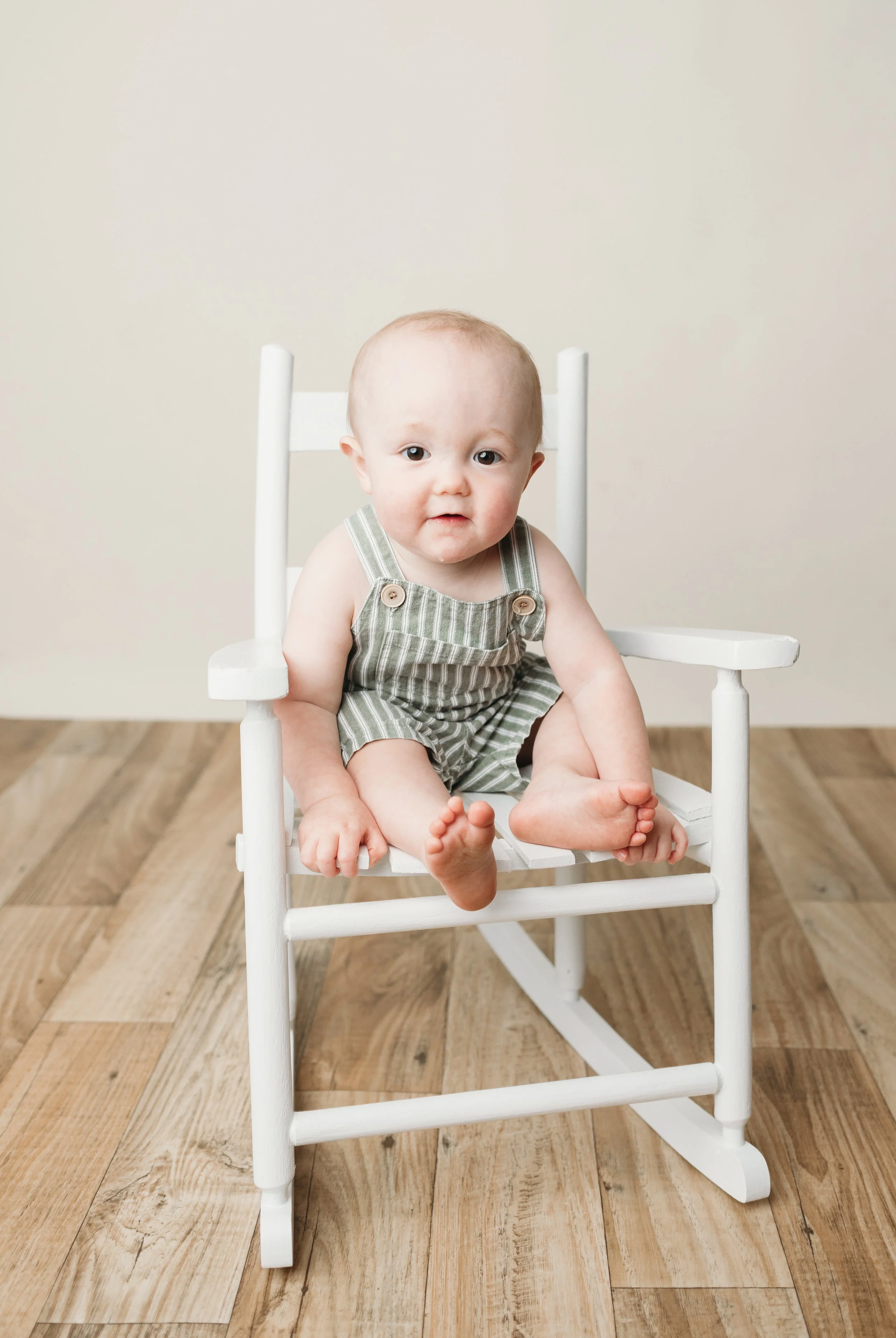 A milestone session with our white wooden rocking chair,  baby is wearing a green and white striped outfit, on a wooden floor with a plain beige background.