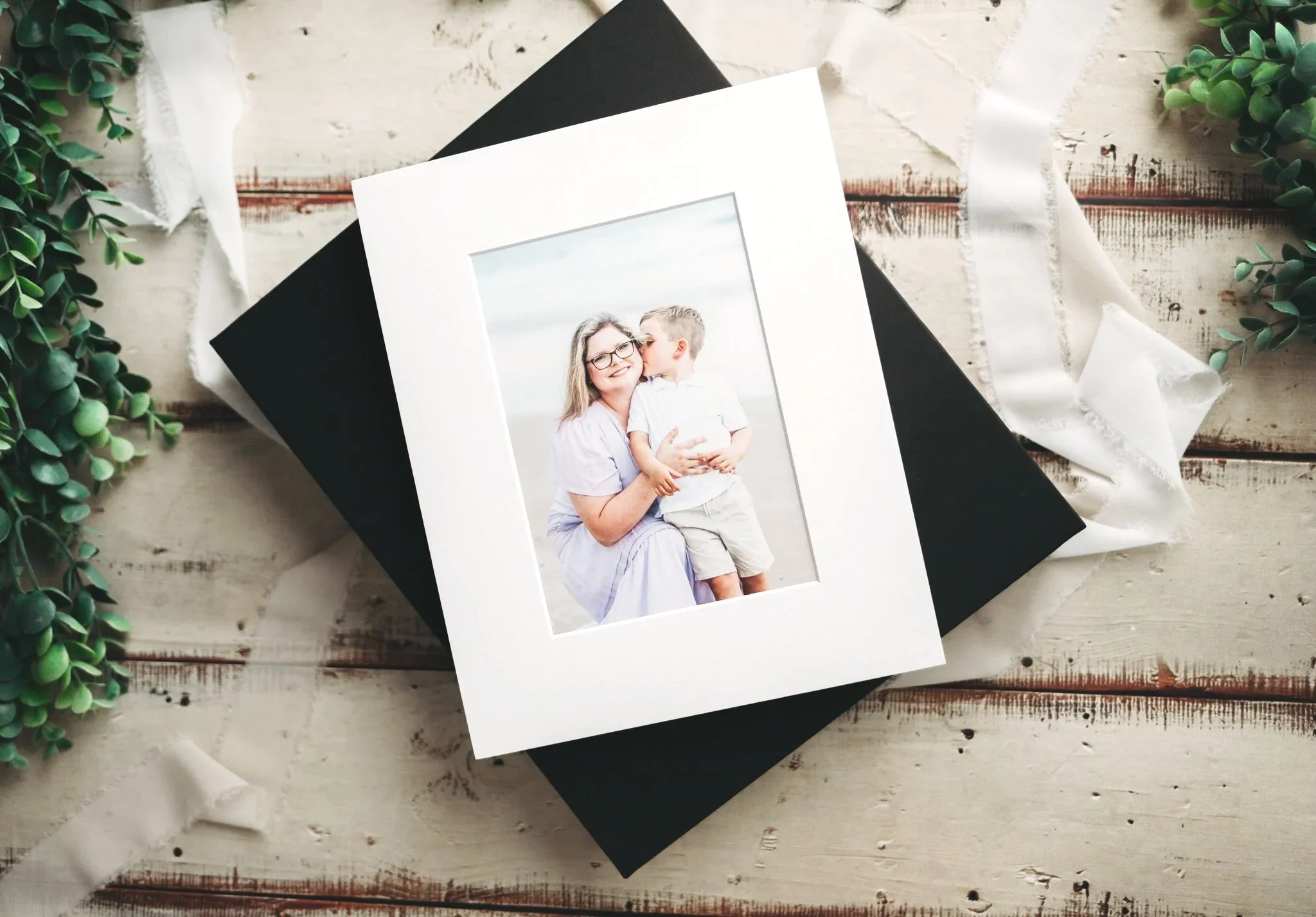 A framed photograph of a woman and a young boy, with the woman smiling and the boy giving her a kiss, placed on top of two closed books, surrounded by greenery and white ribbons on a rustic wooden surface.