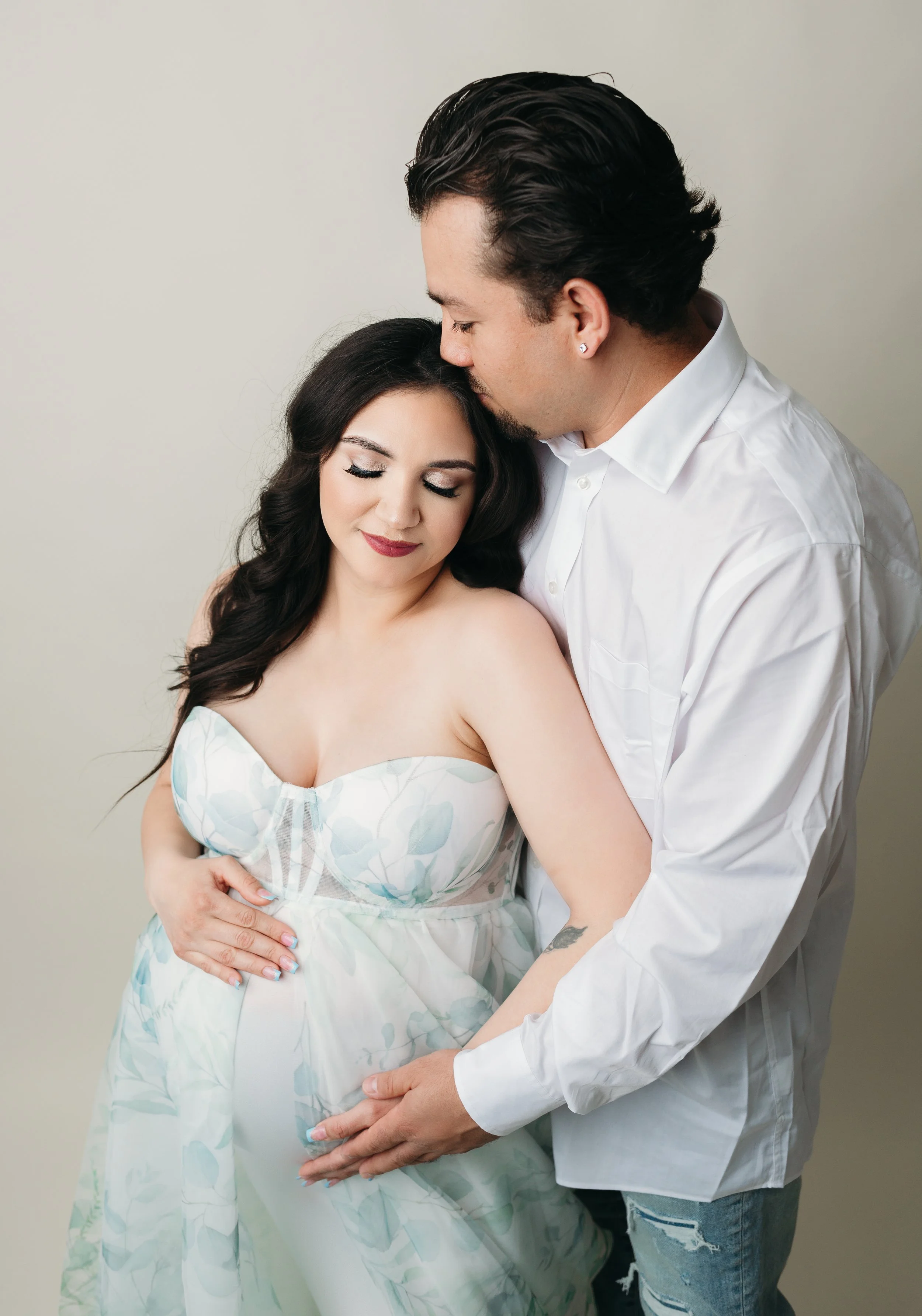 A maternity mama, wearing a strapless patterned dress, is holding her belly. A man with dark hair, wearing a white shirt and jeans, is standing behind her, gently holding her belly and leaning in close, with his eyes closed.