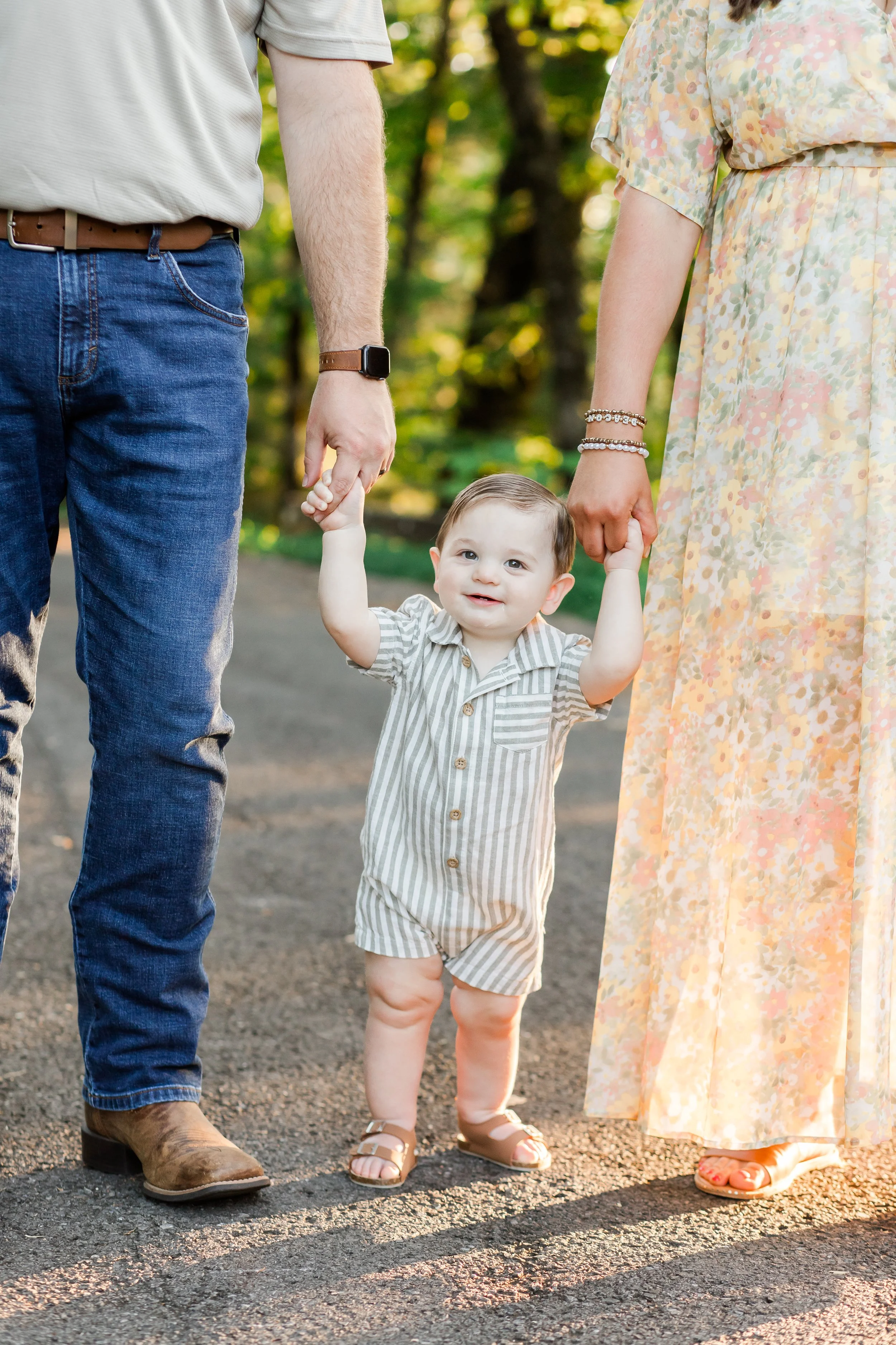 A young child in striped overalls holding hands with two adults, one on each side, walking outdoors in a park or wooded area.