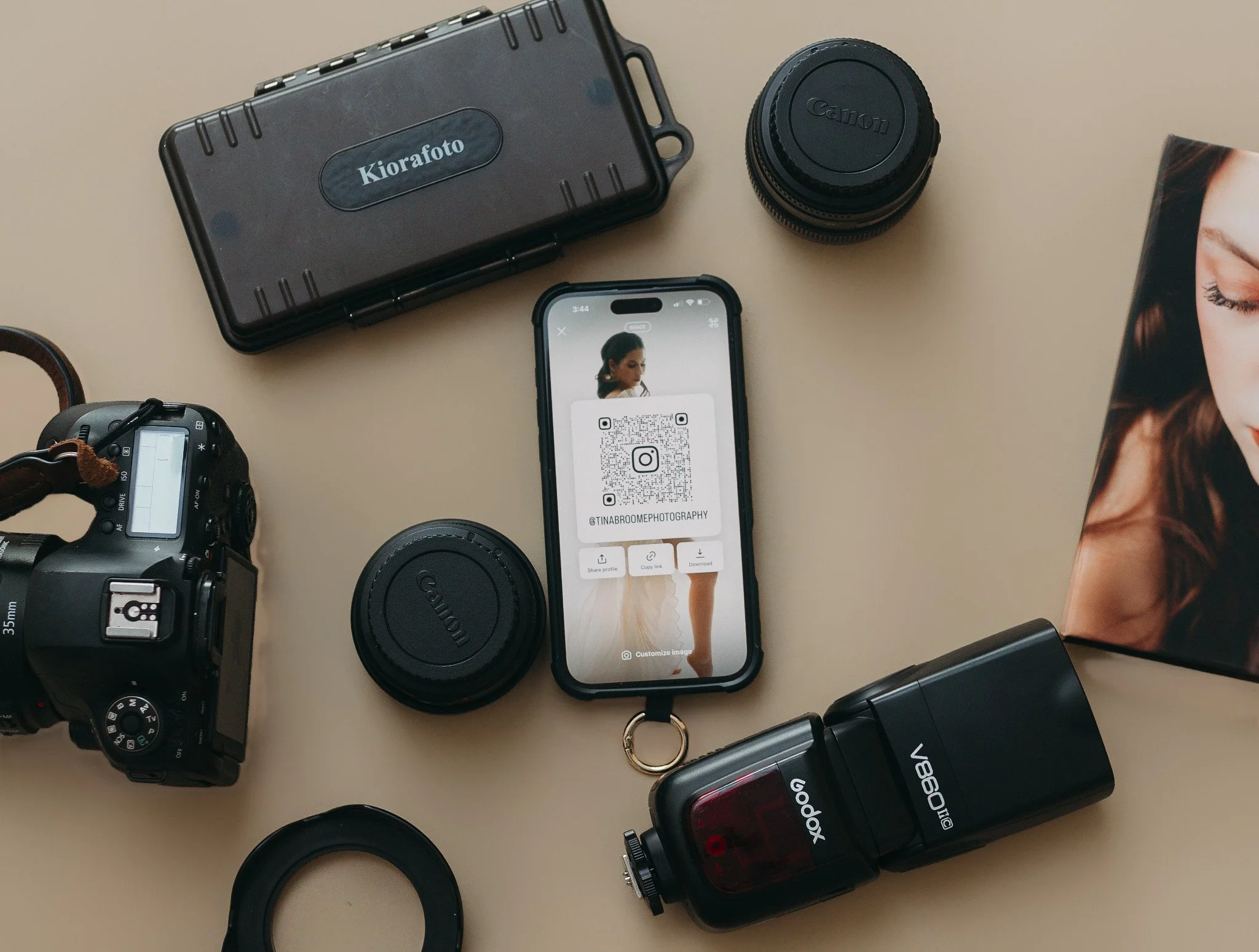 Photography and camera equipment on a beige surface, including a DSLR camera, two camera lenses, a camera flash, a printed photograph, and a smartphone displaying a QR code for photography branding.