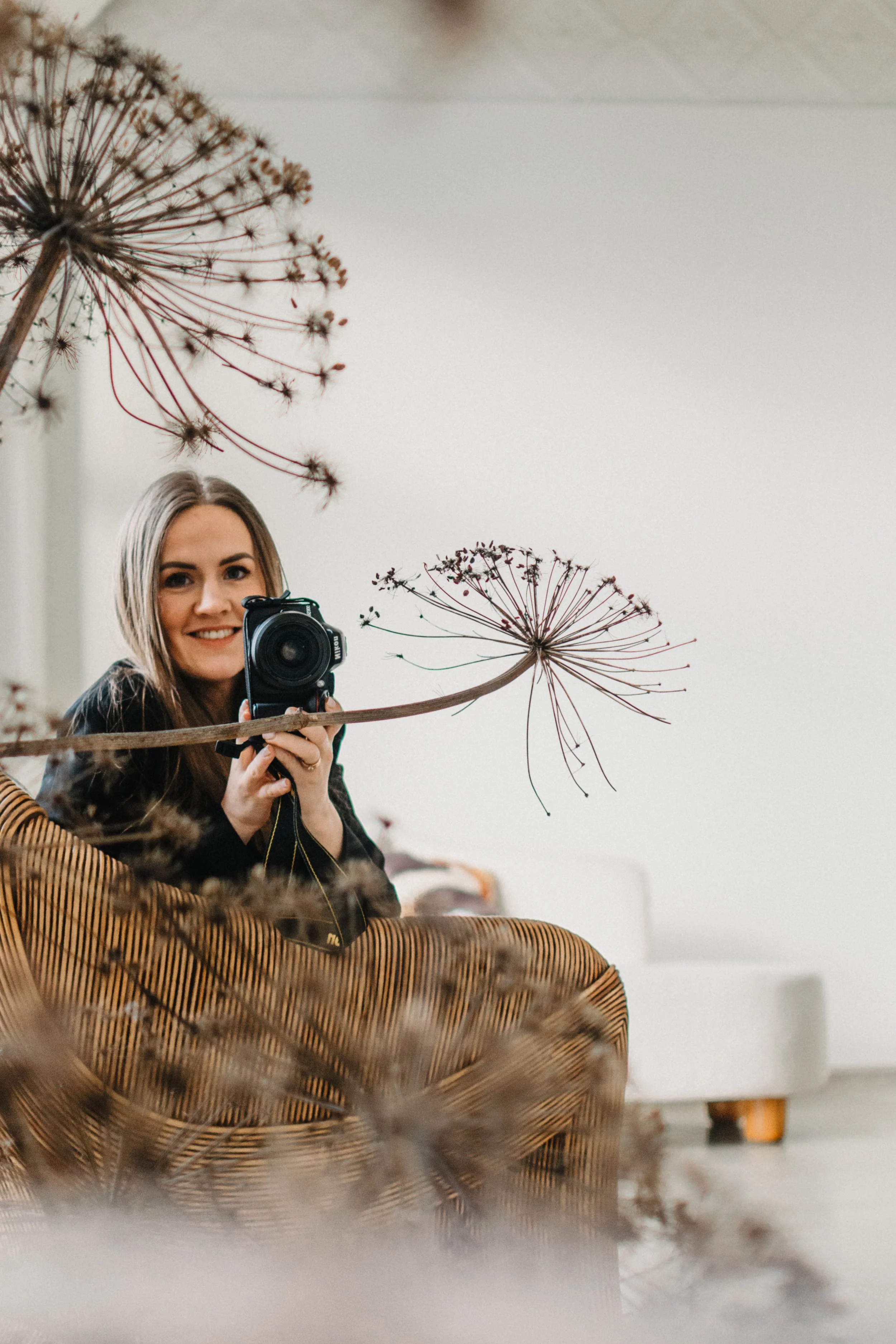 A woman taking a selfie with a camera in front of her, sitting on a patterned chair, surrounded by dried plant stems and seed heads in an indoor setting with a plain background.