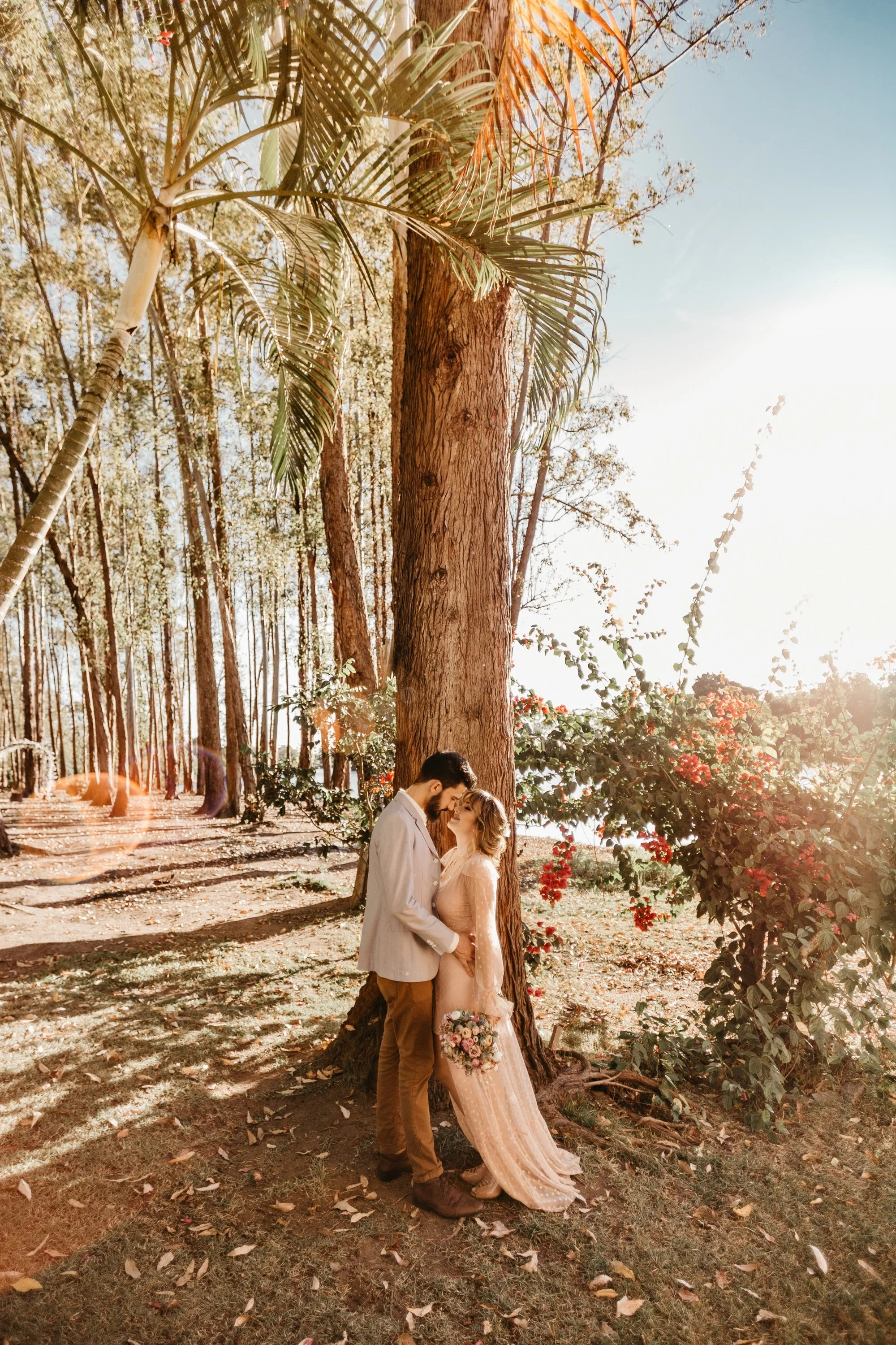 A couple in wedding attire embracing under a large tree in a sunlit outdoor setting with trees and bushes, holding a bouquet of flowers.