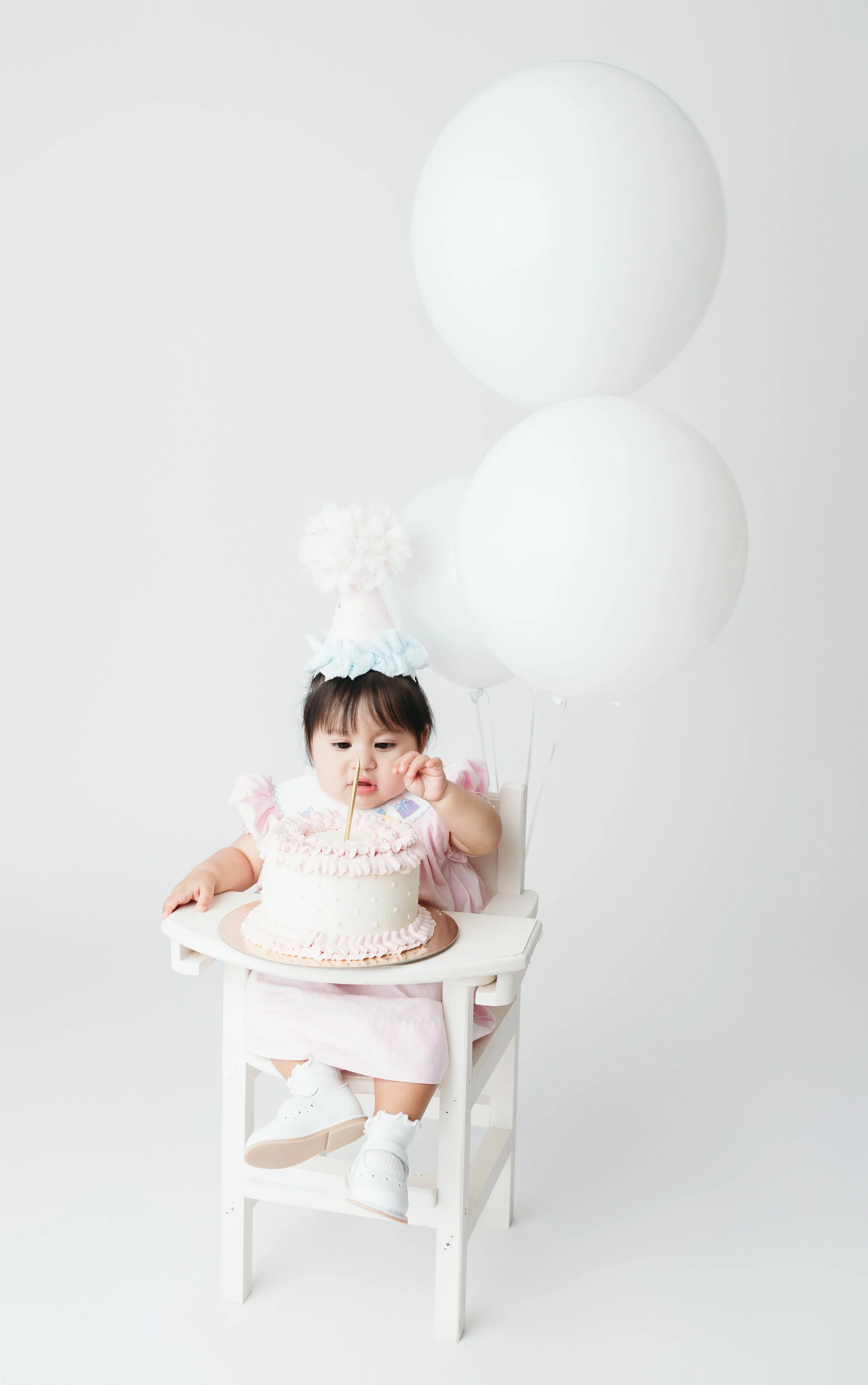 A young girl in a pink dress and birthday hat sitting in a high chair with a birthday cake and white balloons in a plain white background. For a cake smash session.