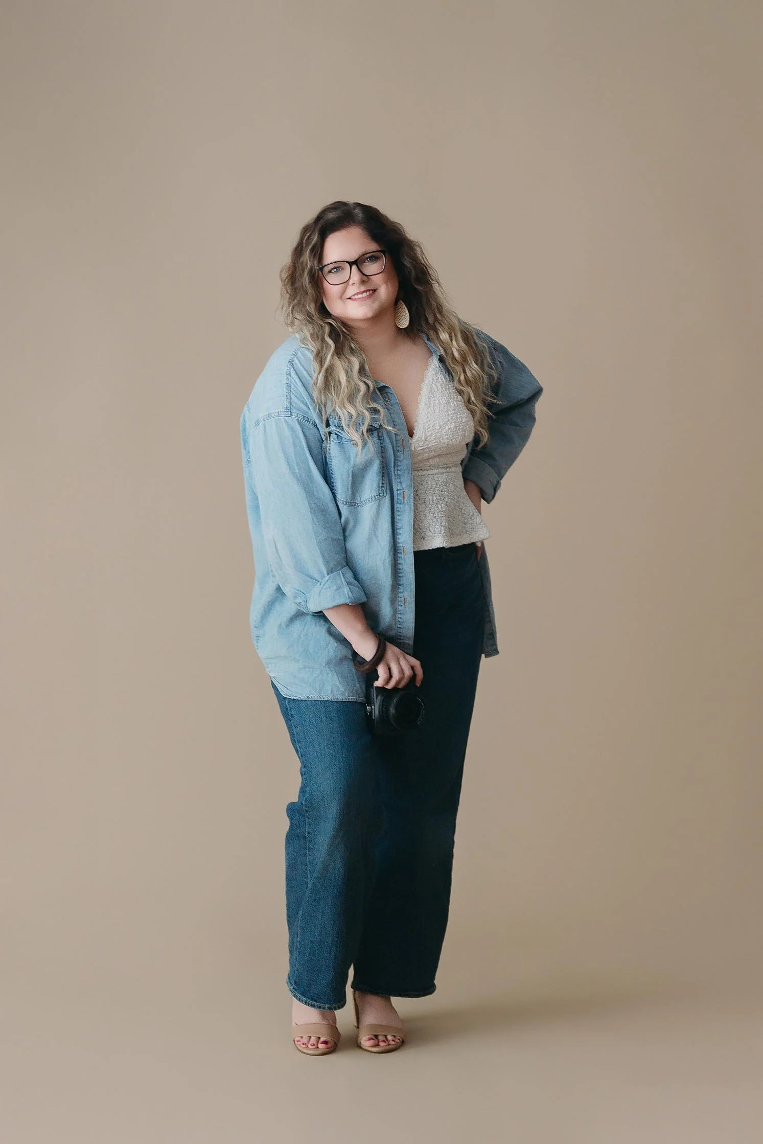 Tina Broome with curly blonde hair, wearing glasses, a white top, a blue denim jacket, dark jeans, and beige heels, holding a camera, standing against a beige background.