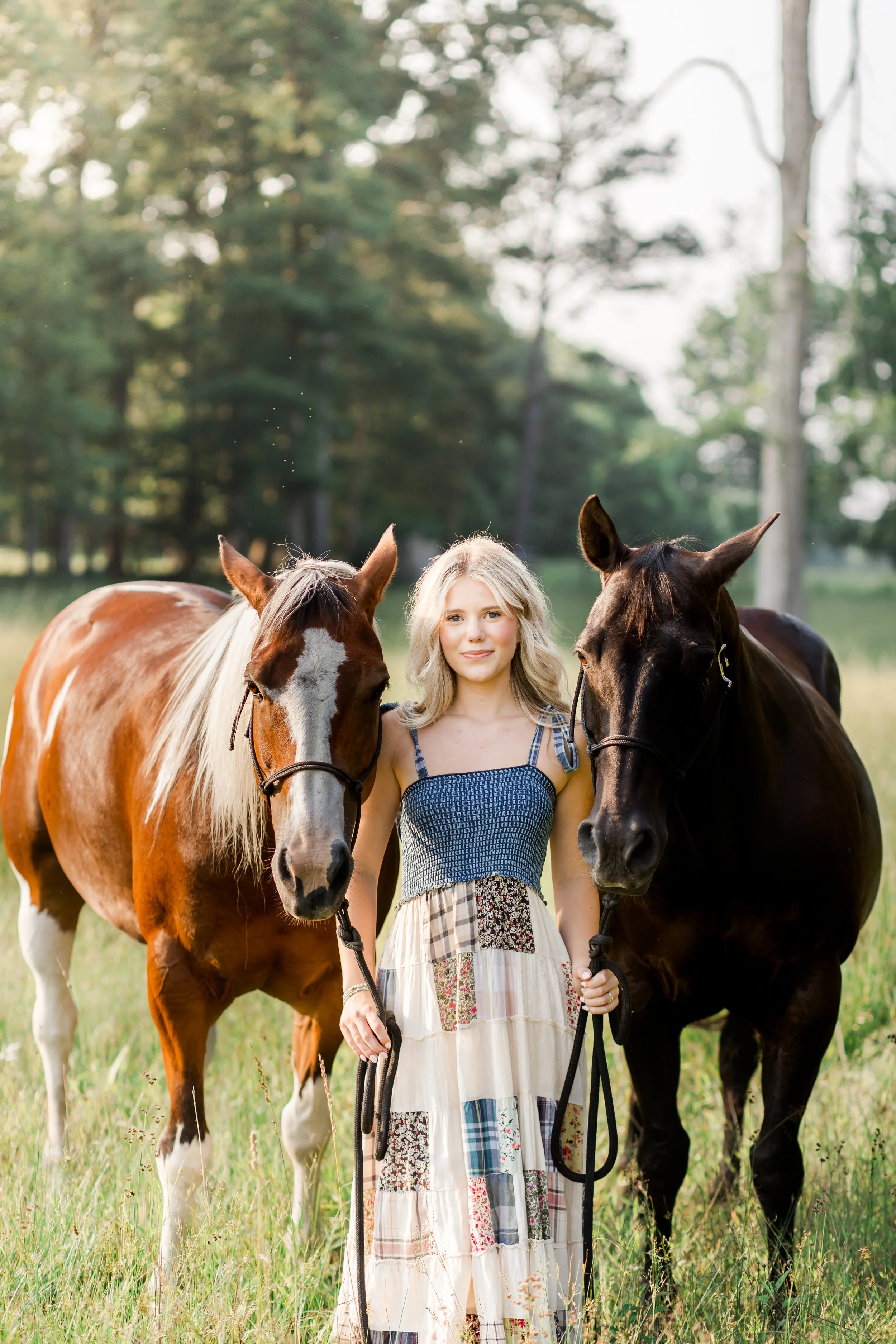 A senior session of Lilly standing between two horses in a grassy field, holding the reins of each horse, with trees in the background.