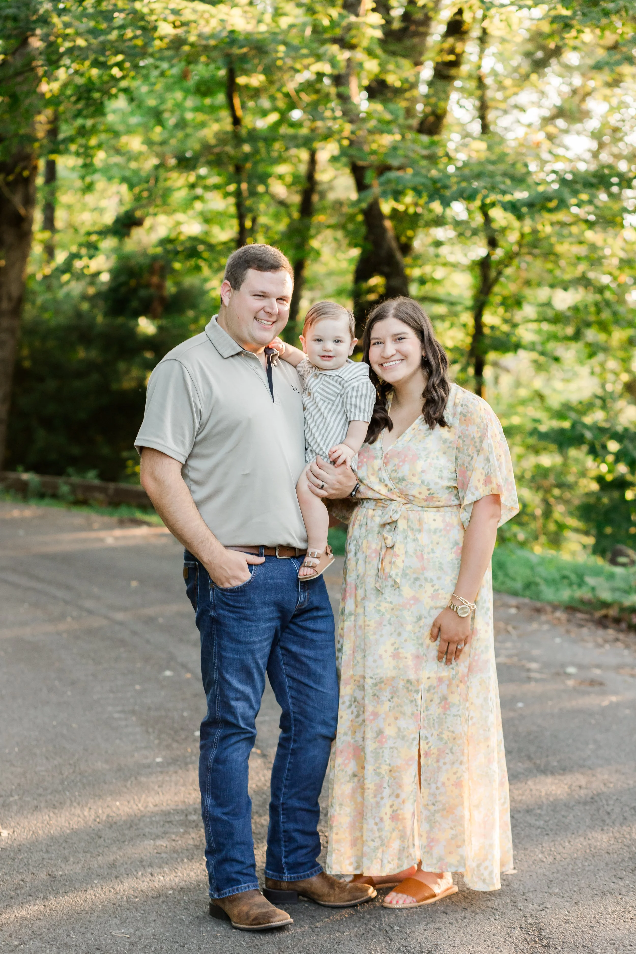 A family of three outdoors, standing on a paved path with green trees in the background. The man is wearing a beige polo shirt and blue jeans, holding a young child in striped clothing. The woman is wearing a long, floral dress. All are smiling.