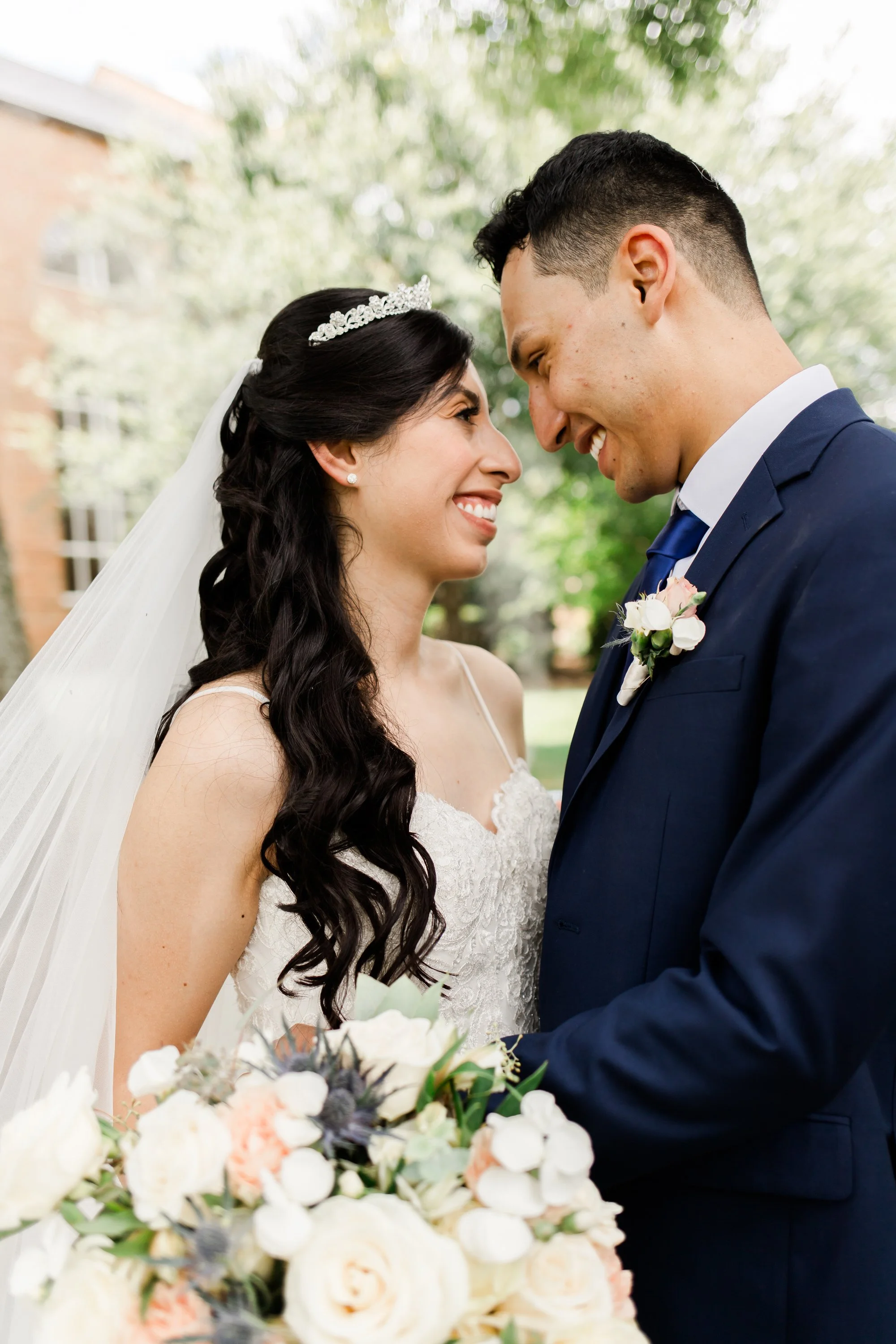 A bride and groom smiling and leaning close to each other outdoors, with the bride holding a large bouquet of white and blush flowers.