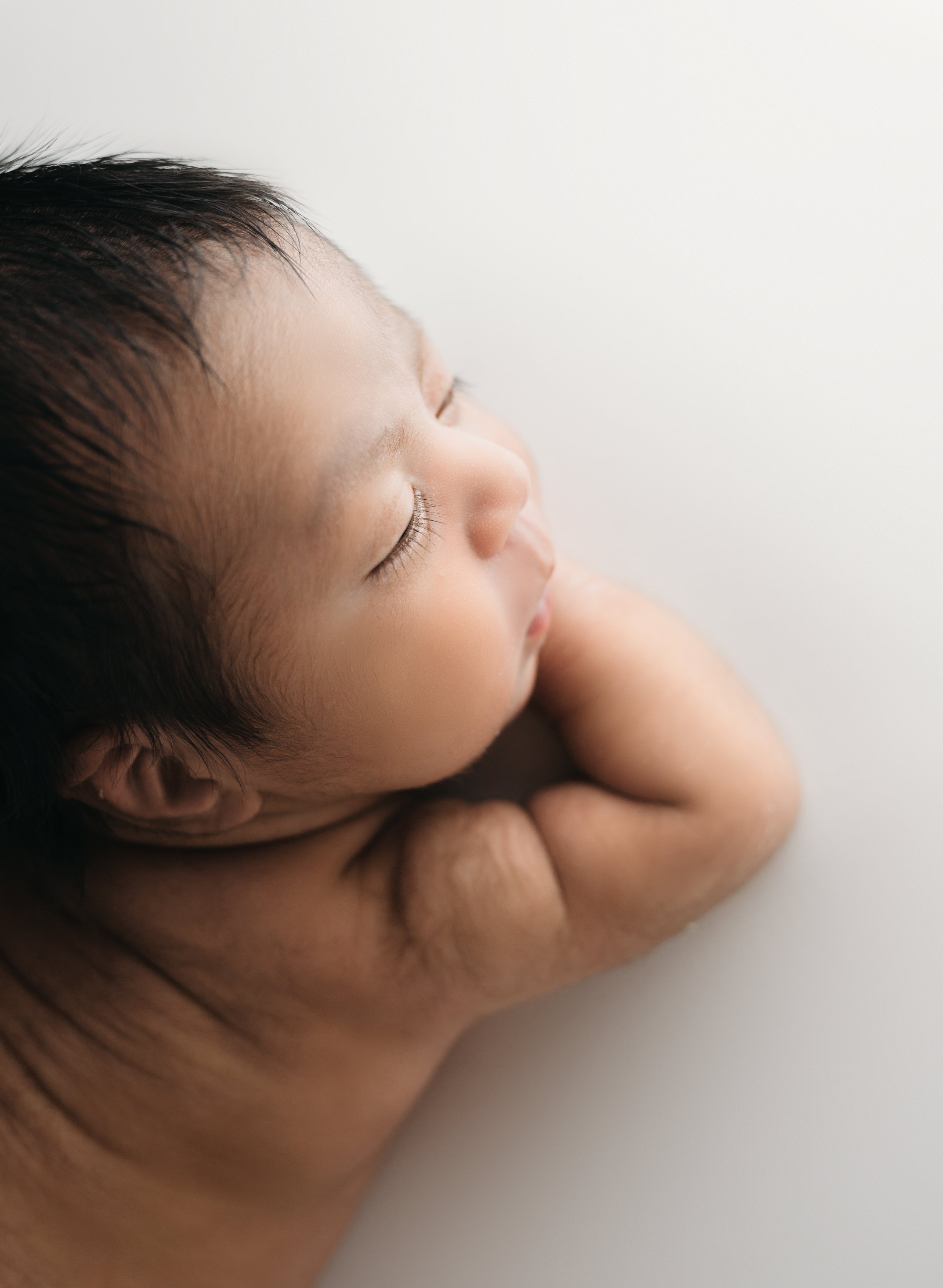 Close-up of a sleeping newborn baby's face with eyes closed, resting on an adult's arm on a white surface.