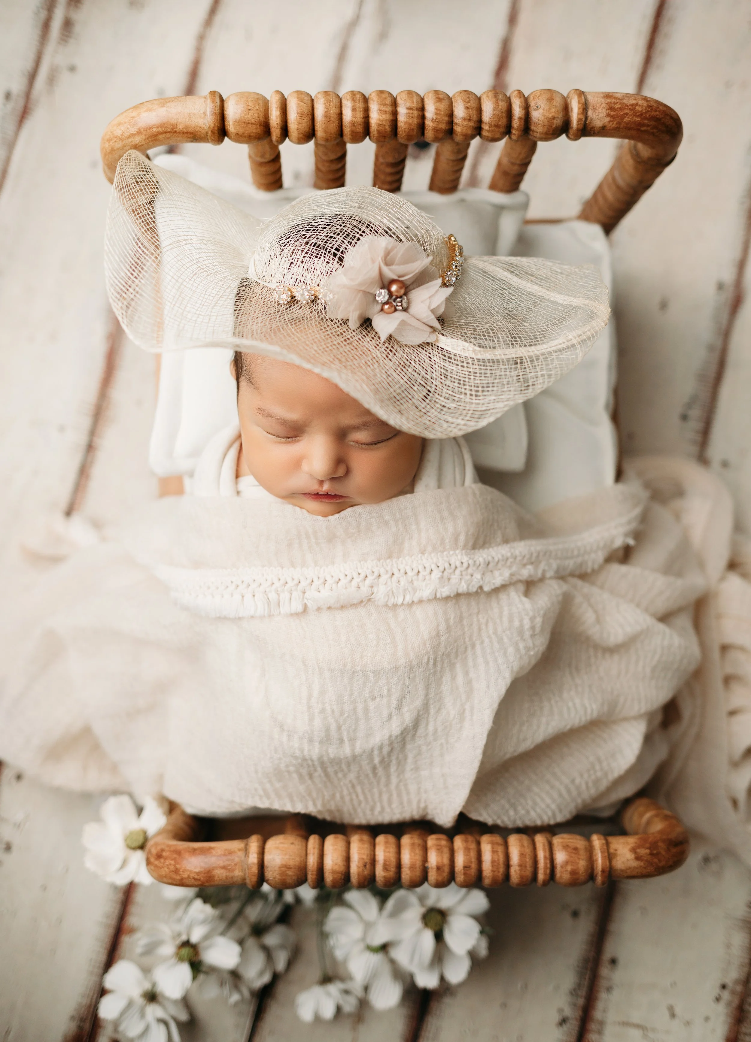 A newborn baby lying in a small wooden bed, wearing a large cream-colored hat with a decorative flower and pearls, wrapped in a cream knit blanket. The bed is decorated with white flowers around it, and the background is a wooden plank floor.